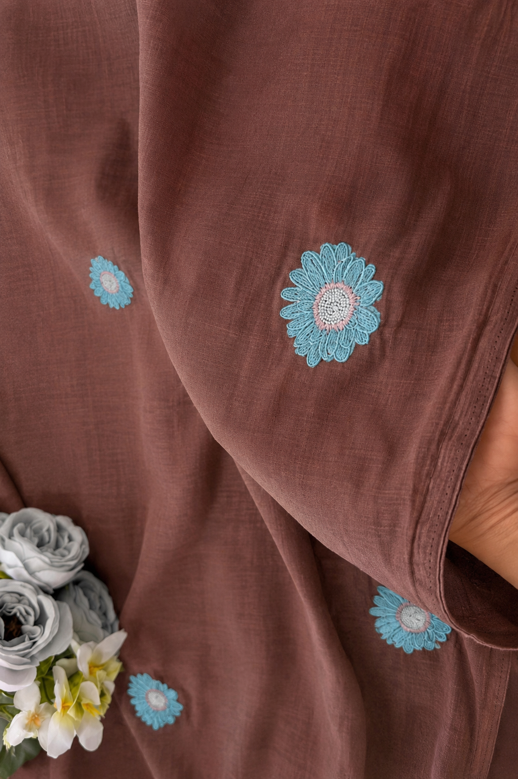 Cocoa brown mul cotton saree draped in soft folds, scattered with aqua daisies embroidered with cream-and-peach centres.