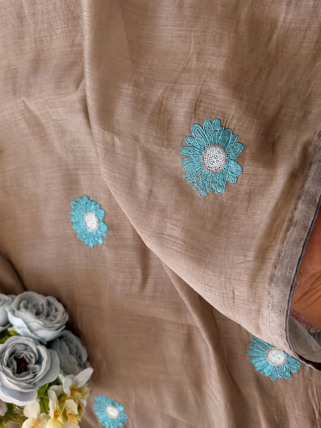 Chiku mul cotton saree with scattered aqua daisy embroidery, styled with soft blue-grey roses.