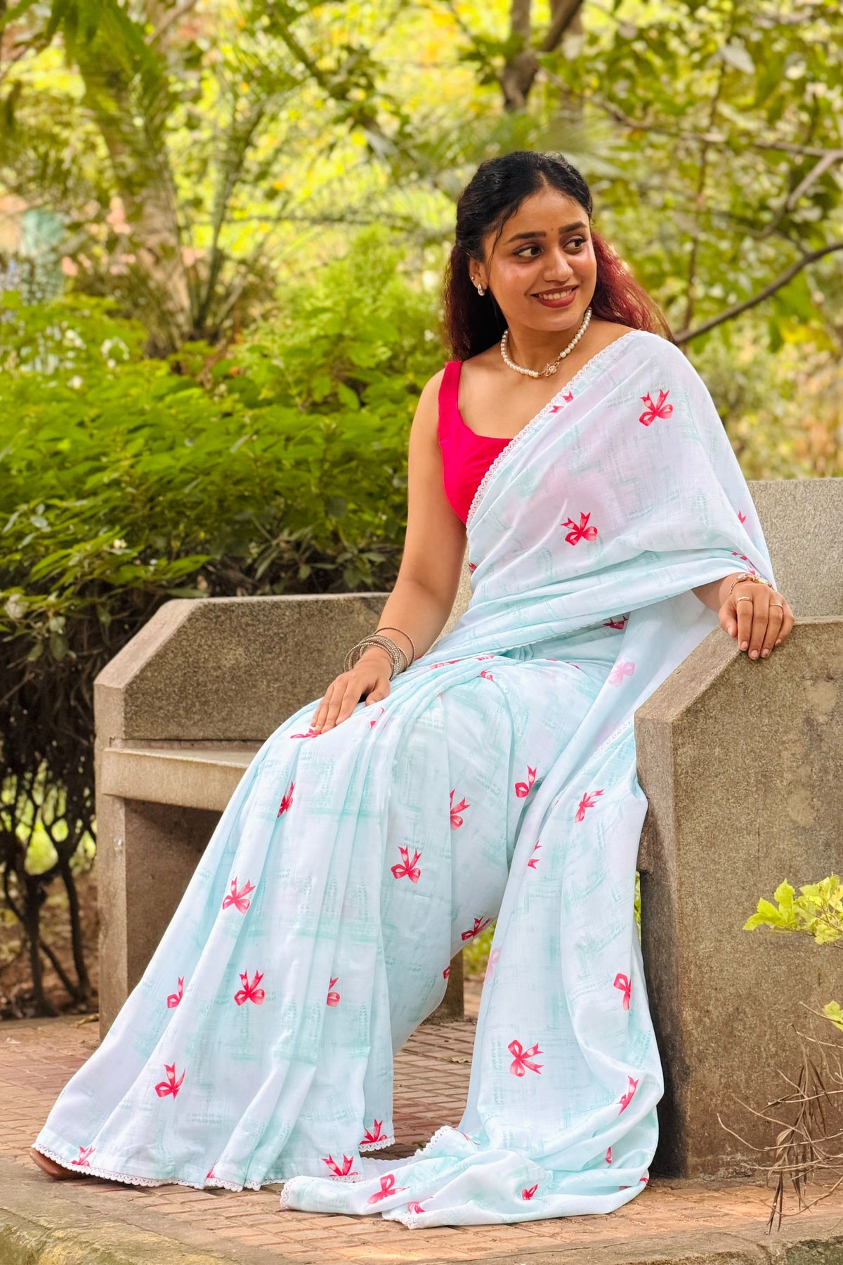 A woman wearing a light blue saree with pink floral embroidery and a bright pink blouse, sitting on a stone bench in a garden setting.