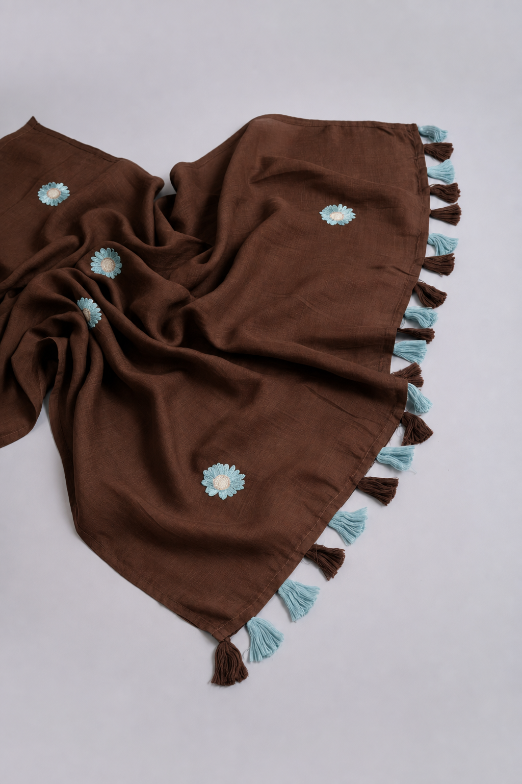 A brown woven scarf with light blue and white embroidered flowers and tassels, laid out on a light gray background.