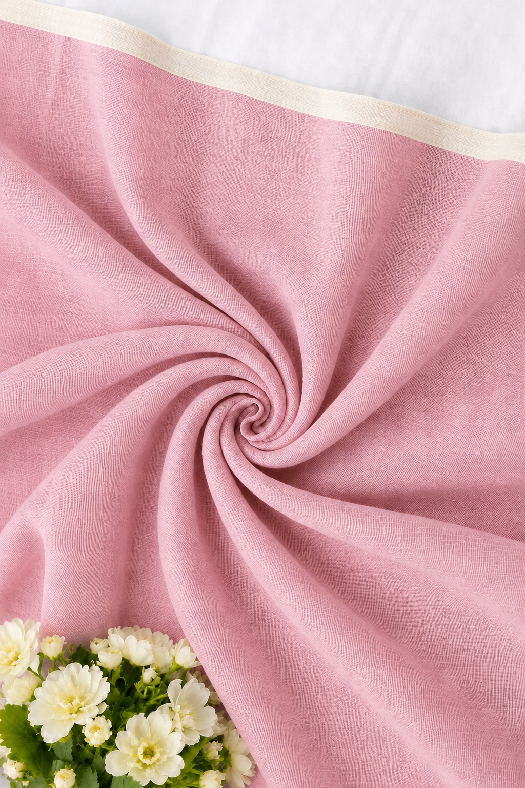 A close-up of a soft pink fabric, likely a throw blanket or scarf, styled in a spiral drape with a cream-colored top border and white flowers in the corner.