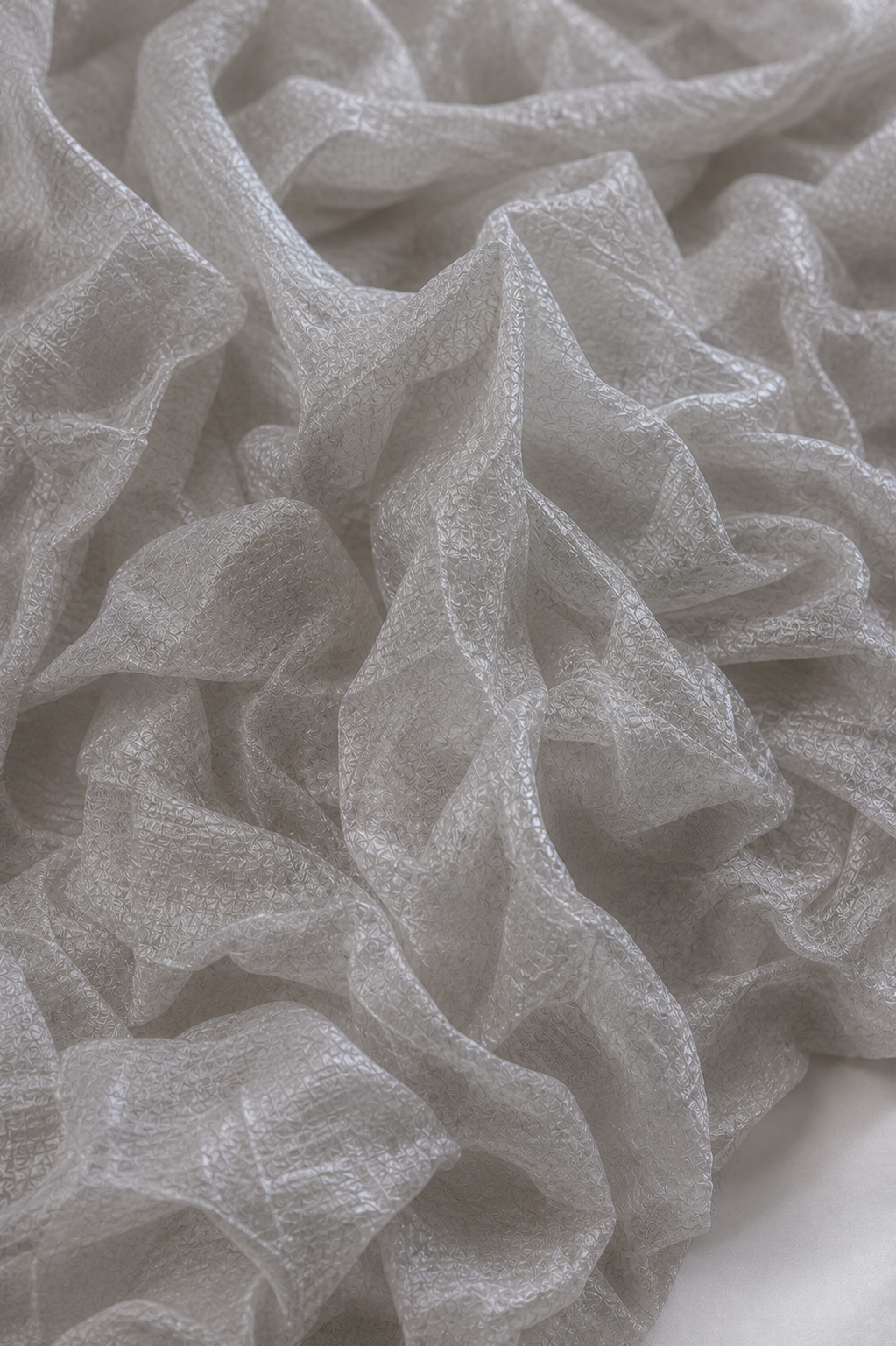 Macro of silver-gray crush tissue in raking light, its irregular crinkle rising in soft ridges and valleys across the sheer weave.