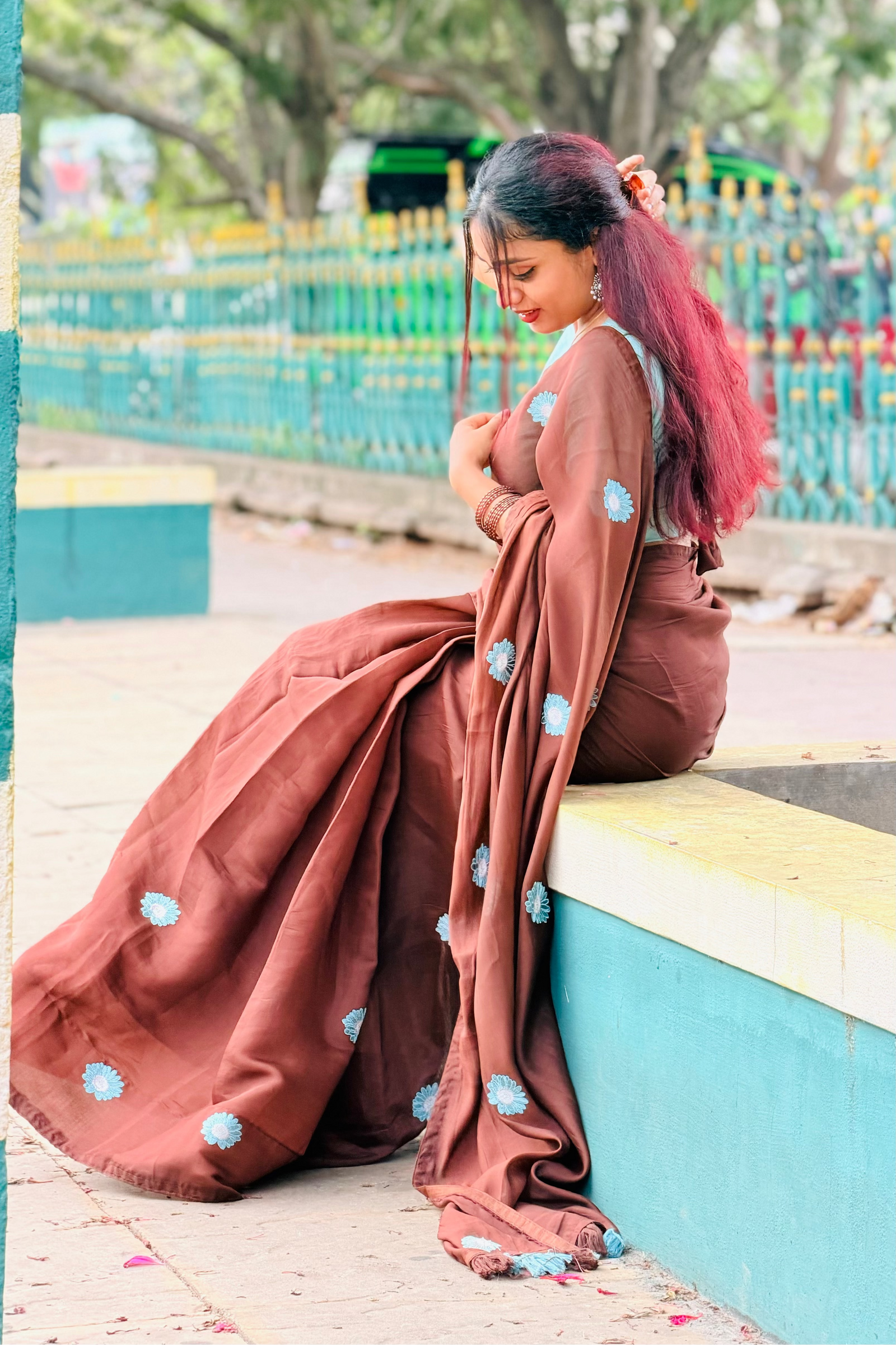 A woman wearing a brown traditional outfit with blue floral patterns sits on a painted concrete ledge outdoors, with trees and a fence in the background.