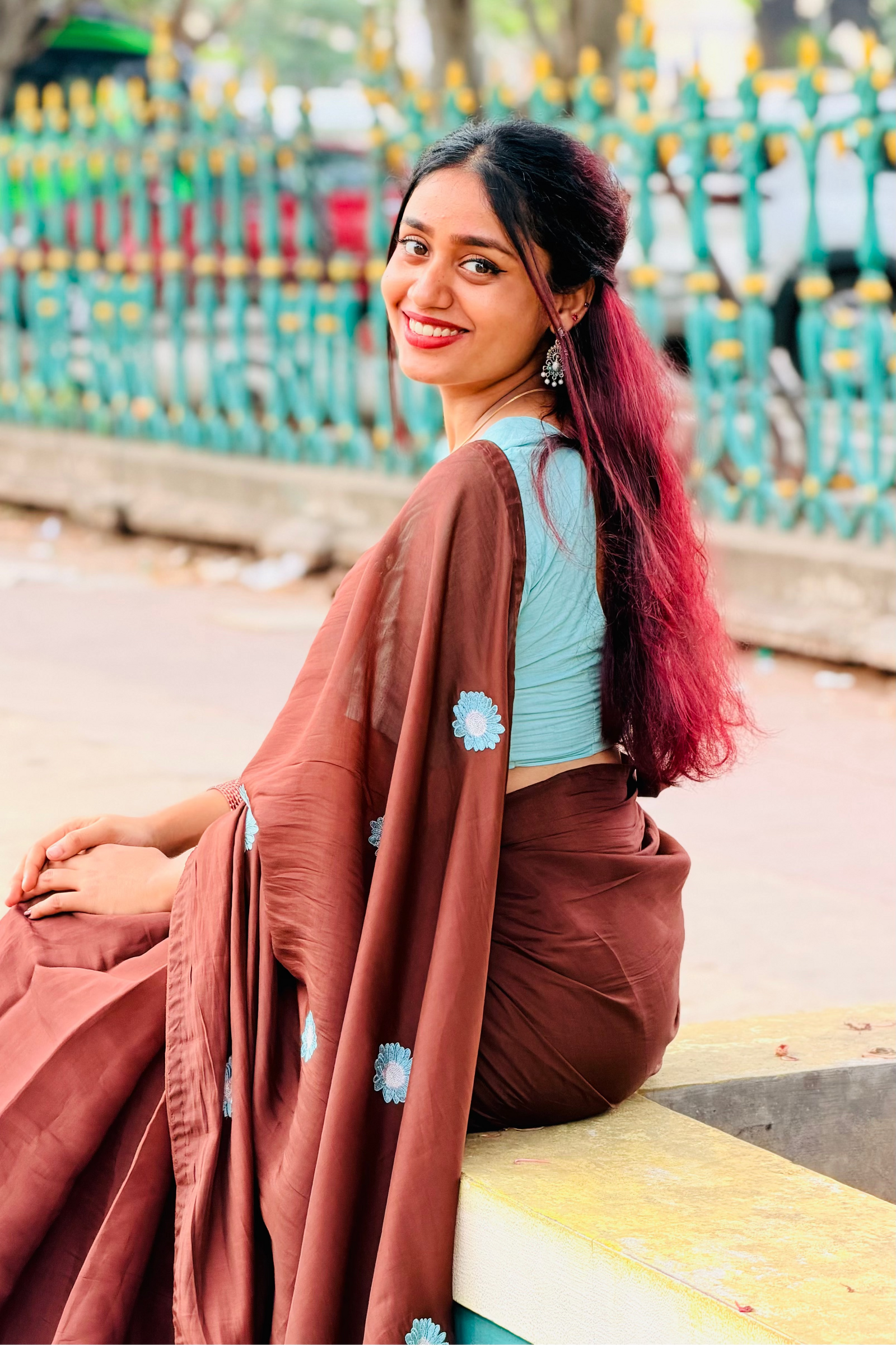 A woman with dark hair featuring pink highlights sits on a yellow curb outdoors, wearing a brown sari with light blue floral embroidery over a light blue crop top.