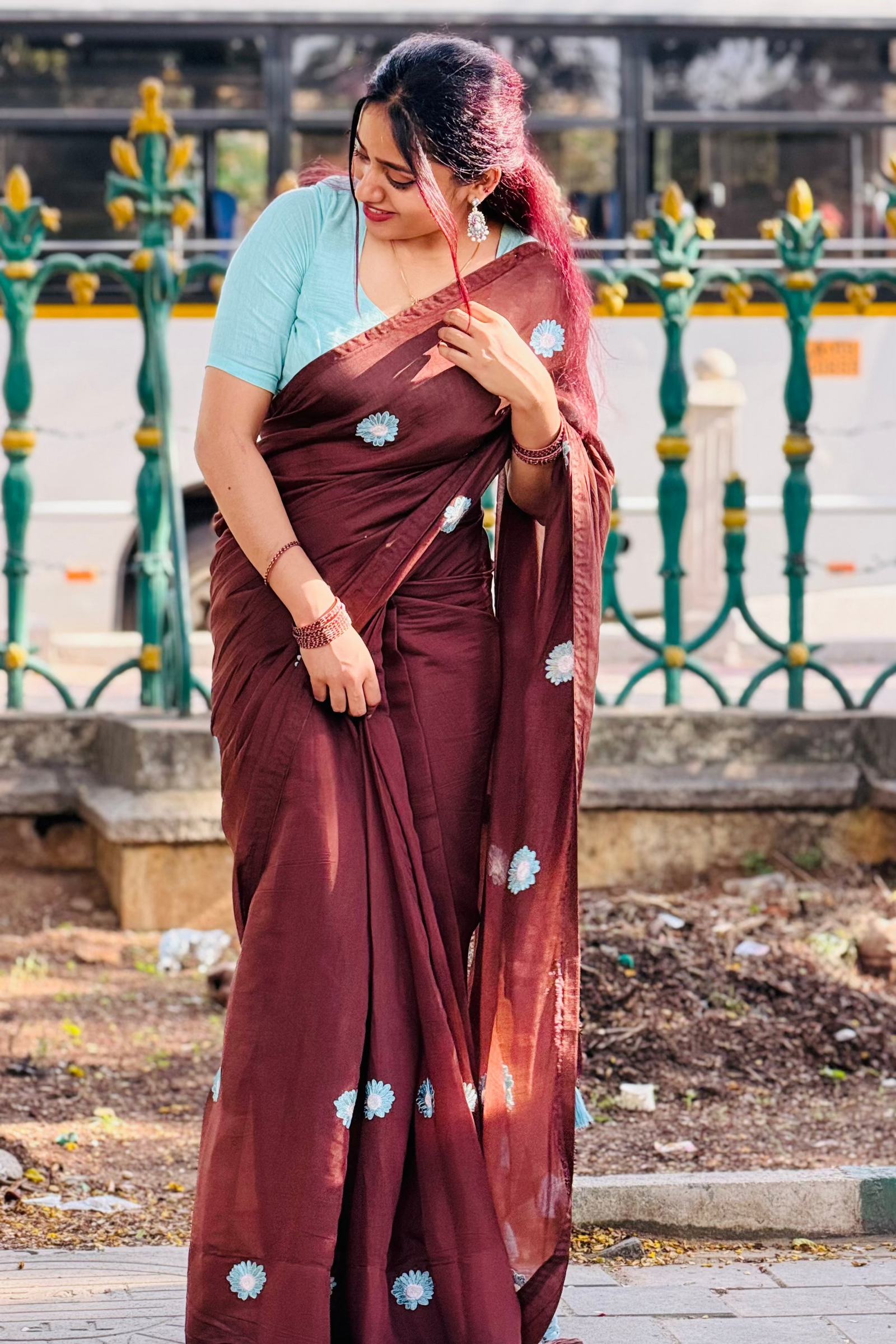 A woman wearing a maroon brown saree with light blue floral prints and a light blue short-sleeve blouse, standing outdoors in front of a green decorative fence.