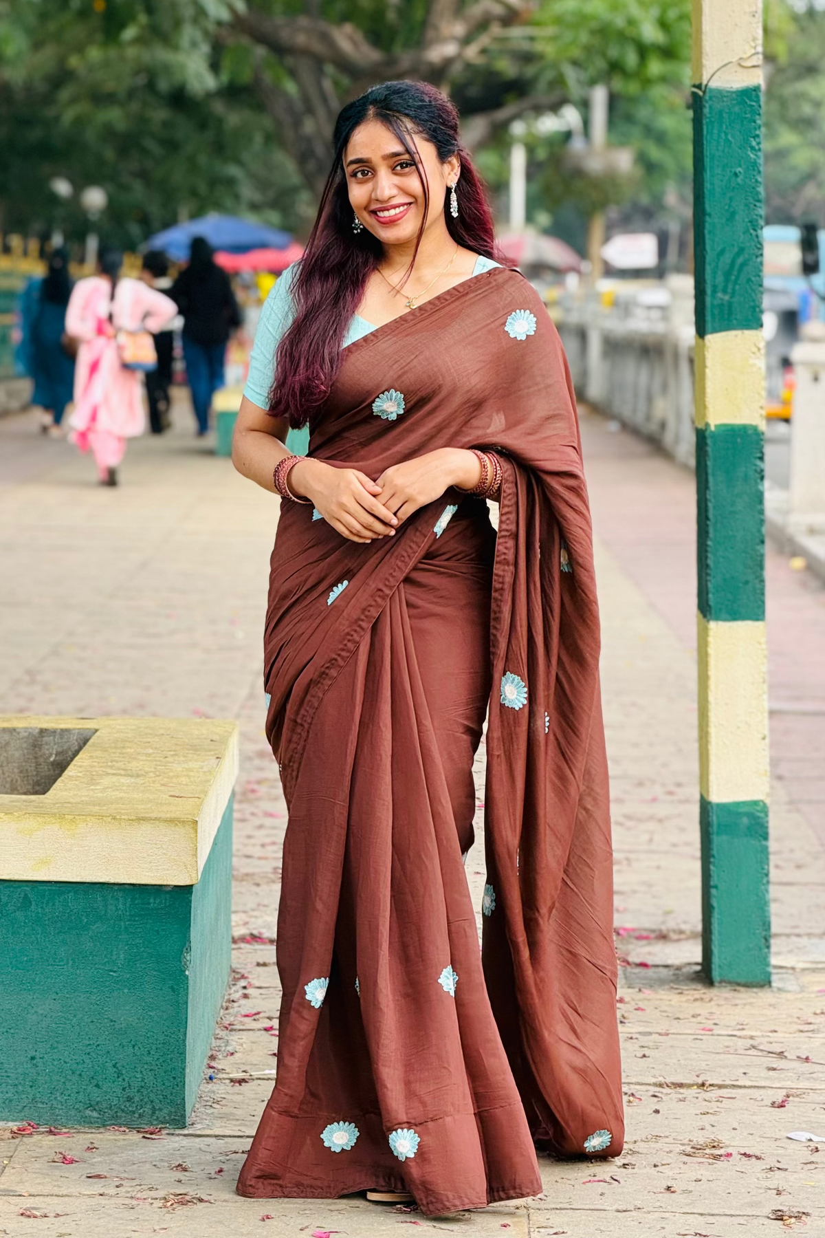 A woman wearing a brown sari with light blue floral motifs and a light blue blouse, standing outdoors on a paved walkway with trees and a green-and-white pole in the background.