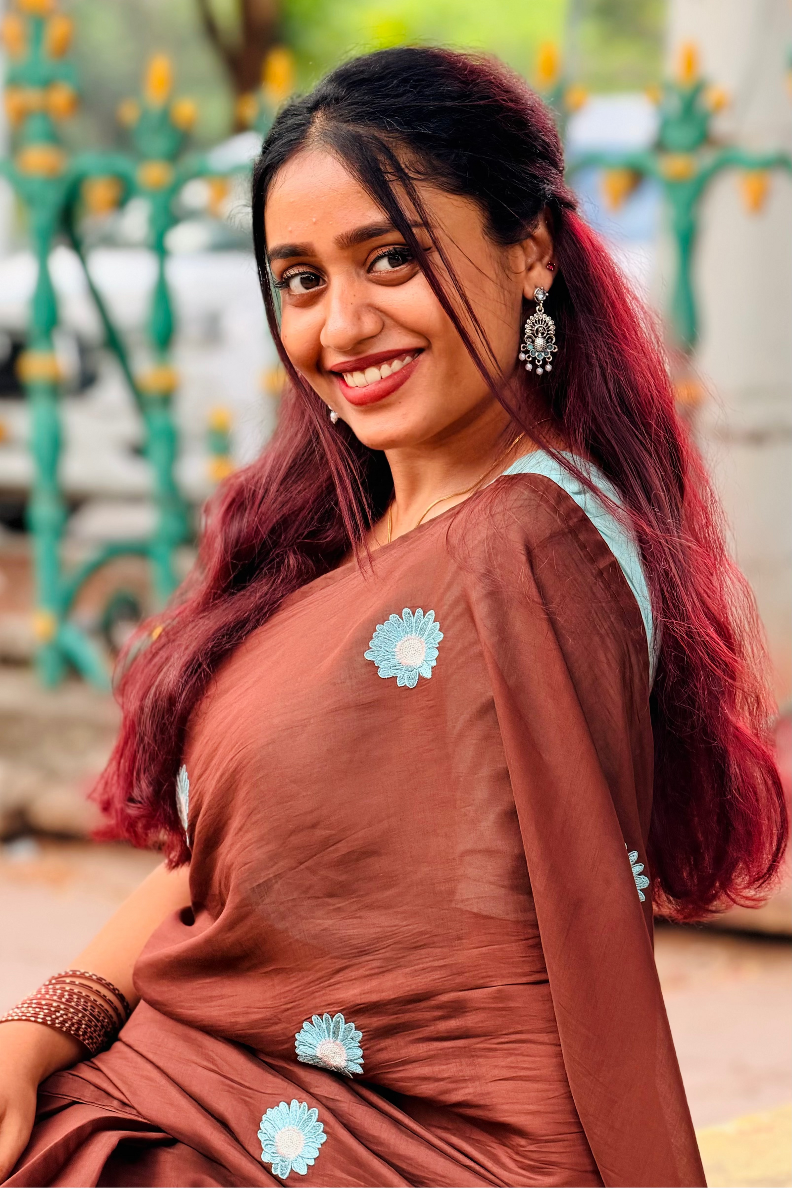 A woman with long dark red hair smiles while wearing a brown sari with light blue floral accents, posed outdoors in front of a blurred green fence.