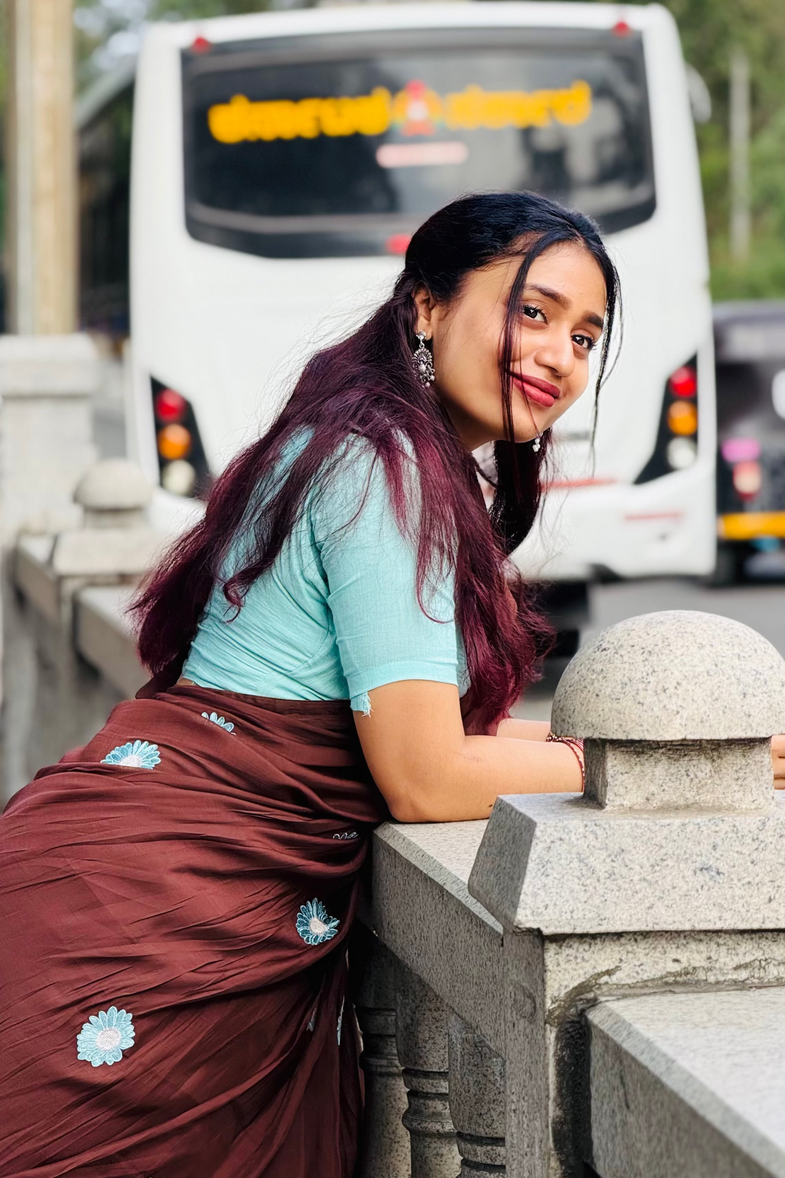 A woman with long dark hair leans on a stone railing, wearing a light blue t-shirt and a maroon sari with small light blue floral motifs. A white bus is blurred in the background.