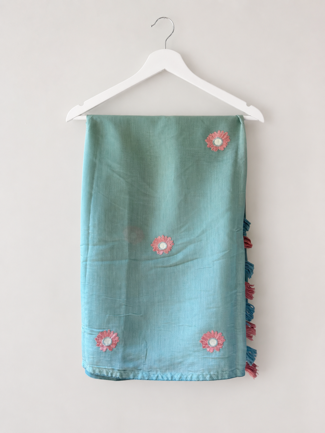 Aqua mul cotton saree with dusty-rose daisy embroidery and pink-and-blue tassels, folded on a hanger.