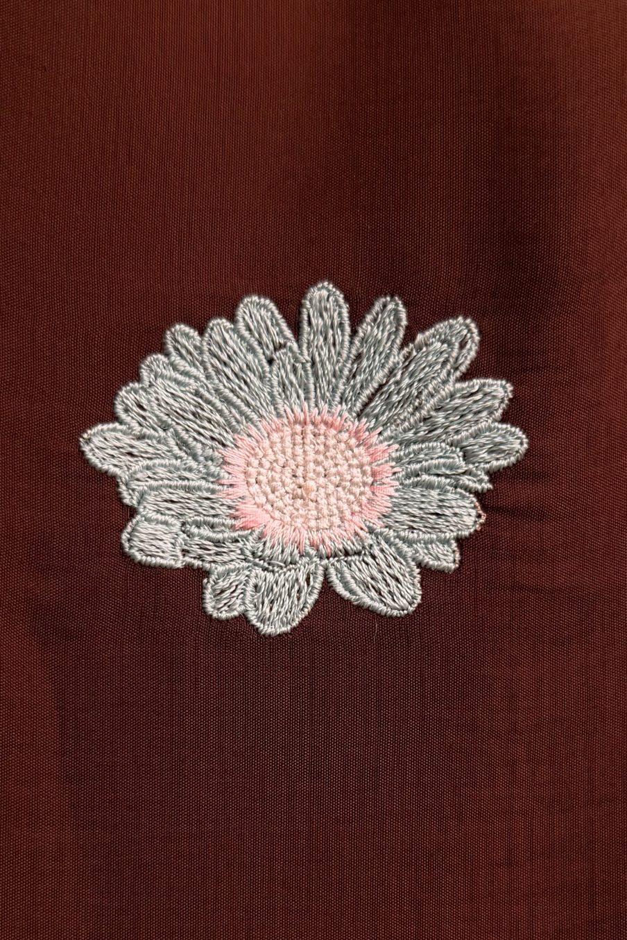 A close-up of a dark brown textured fabric featuring an embroidered daisy flower patch with a light gray stitched petal border and a soft pink center.