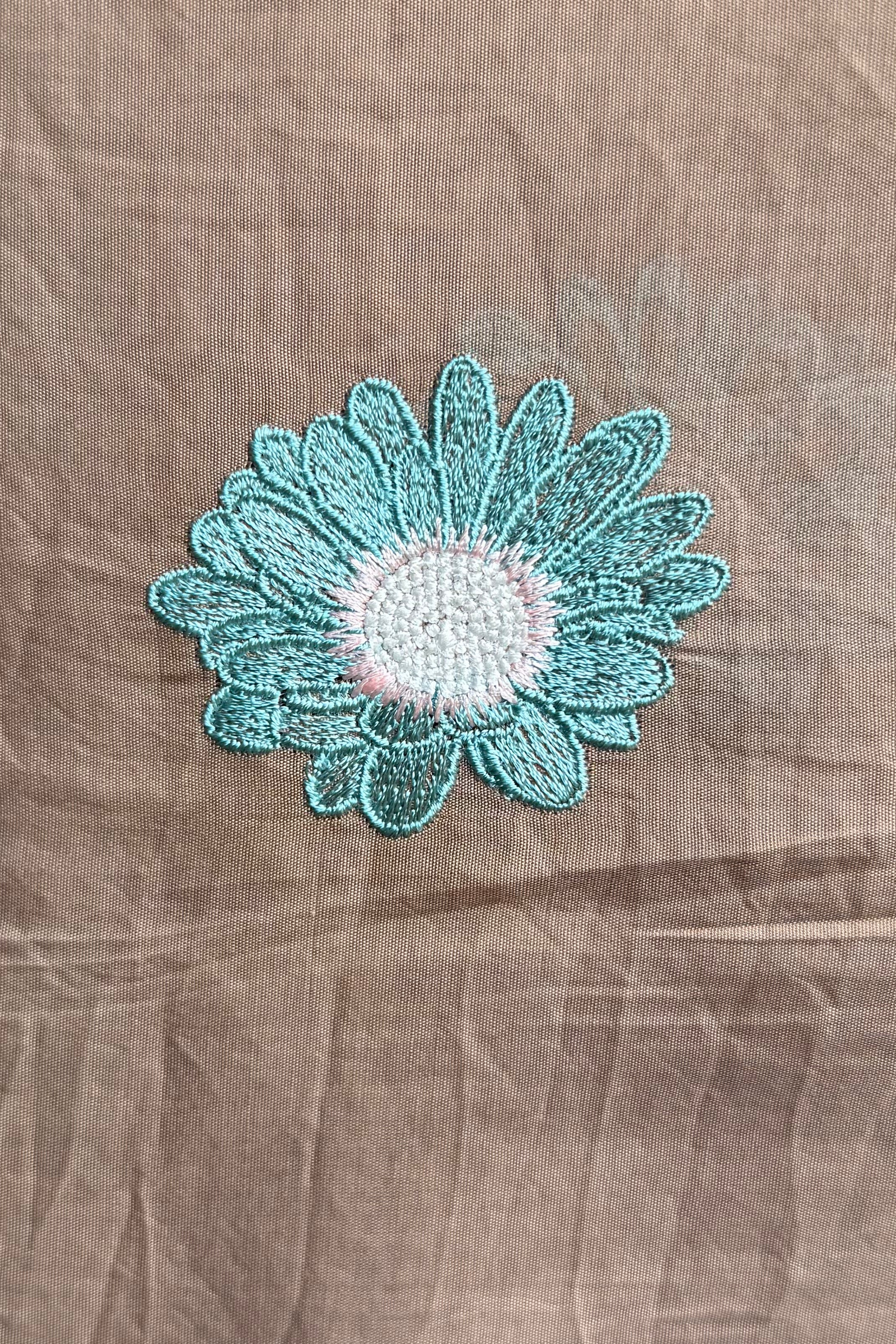 Close-up of a single aqua daisy embroidered in viscose thread on chiku mul cotton.