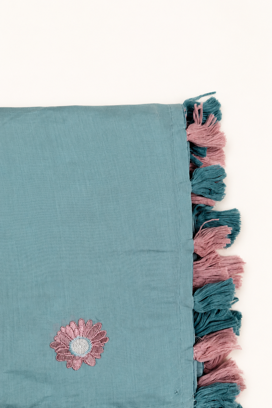 A folded teal blue scarf with pink and teal tassels on one edge and a pink embroidered flower on the front, shown against a plain off-white background.