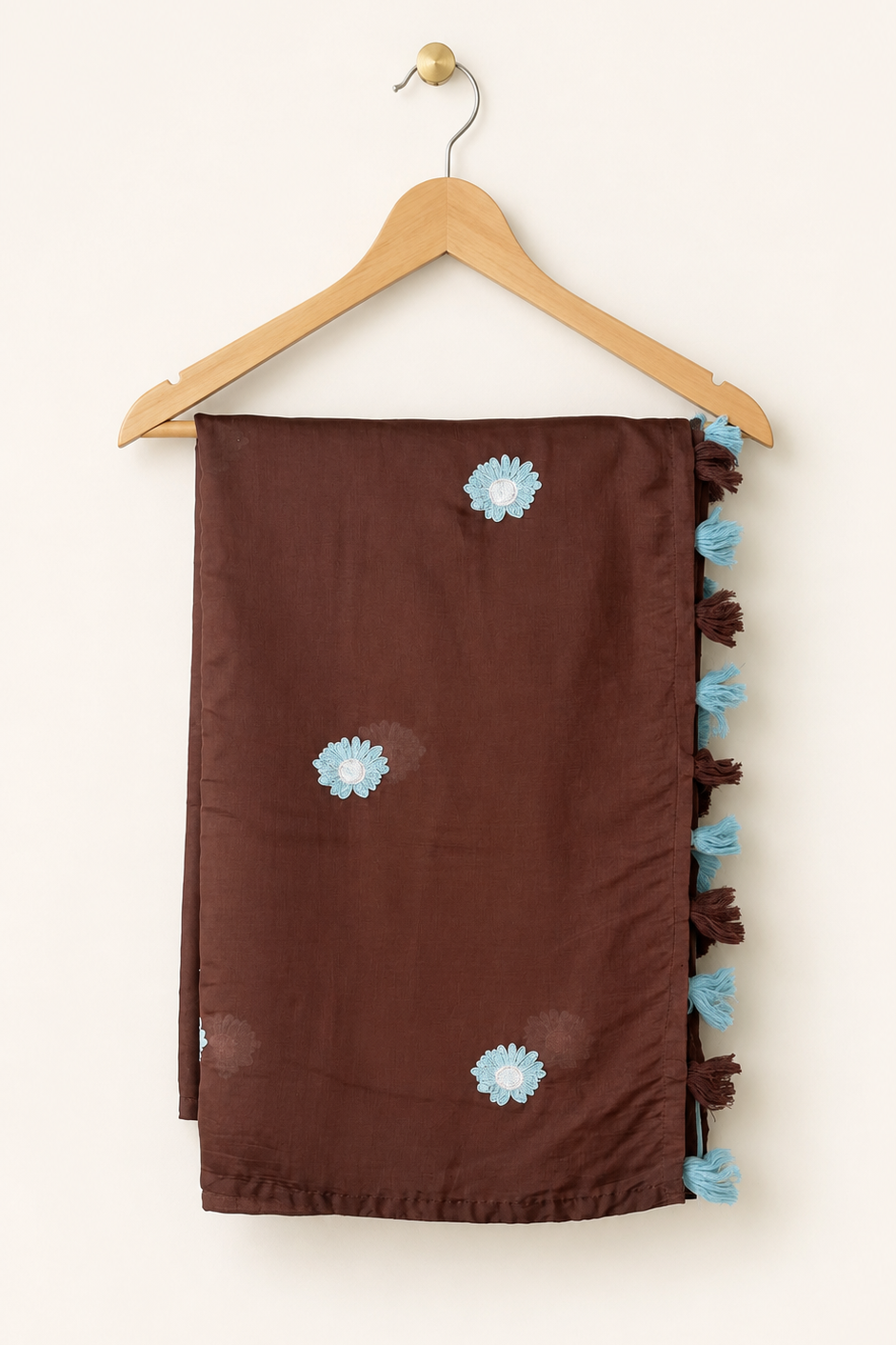 A dark brown scarf with light blue embroidered flowers and a matching tassel trim, hanging on a wooden hanger against a plain off-white background.
