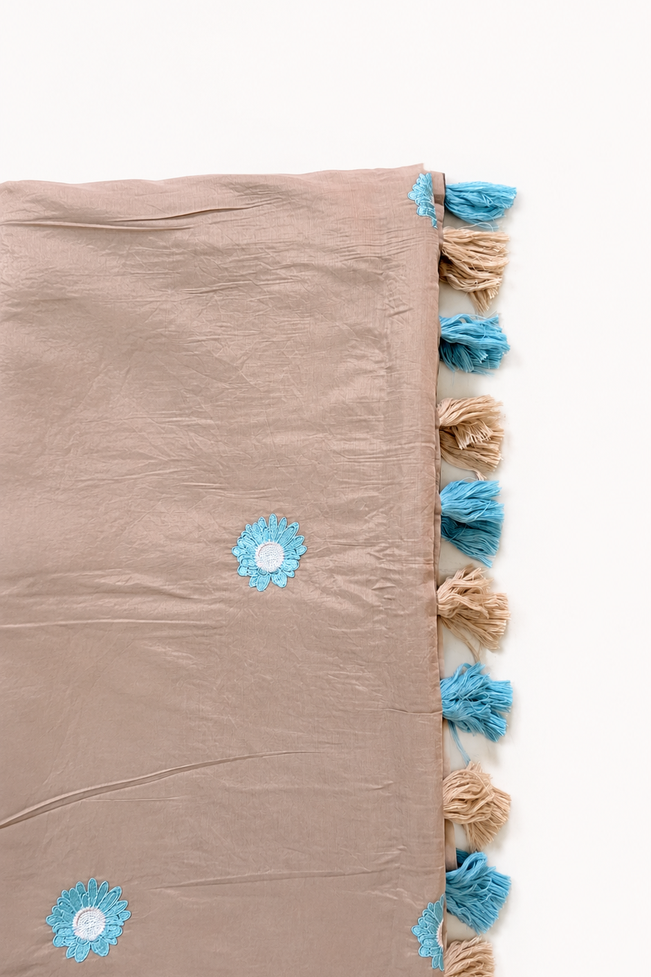 A dusty pink fabric throw blanket with blue and beige tassels along one edge and small blue embroidered flowers, laid flat on a white background.