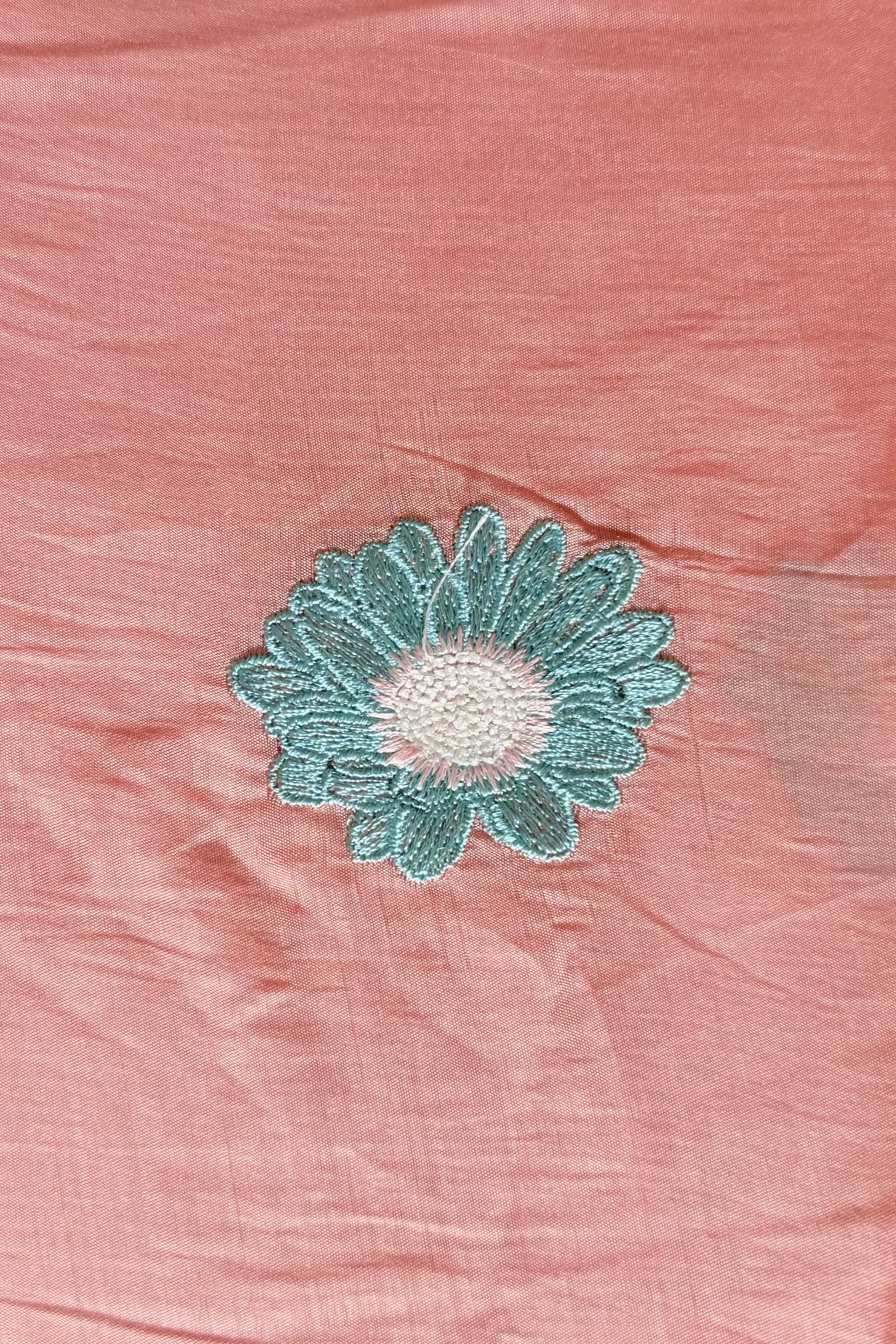 Close-up of blush mul cotton with a single embroidered aqua daisy, a hand resting at the fabric's edge.