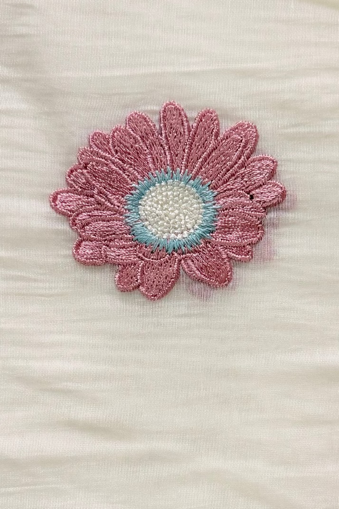 Close-up of a single dusty-rose daisy embroidered in viscose thread, with an aqua ring and cream centre, on ivory mul cotton.