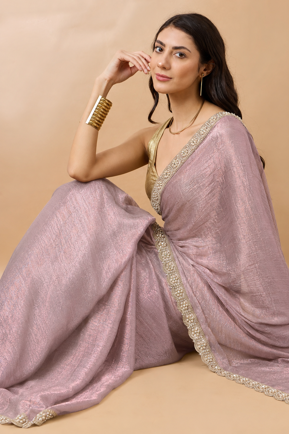 A woman models a lavender-purple saree with gold zari embroidery on a solid beige background. She is wearing a gold blouse, gold bangles, and a gold necklace.