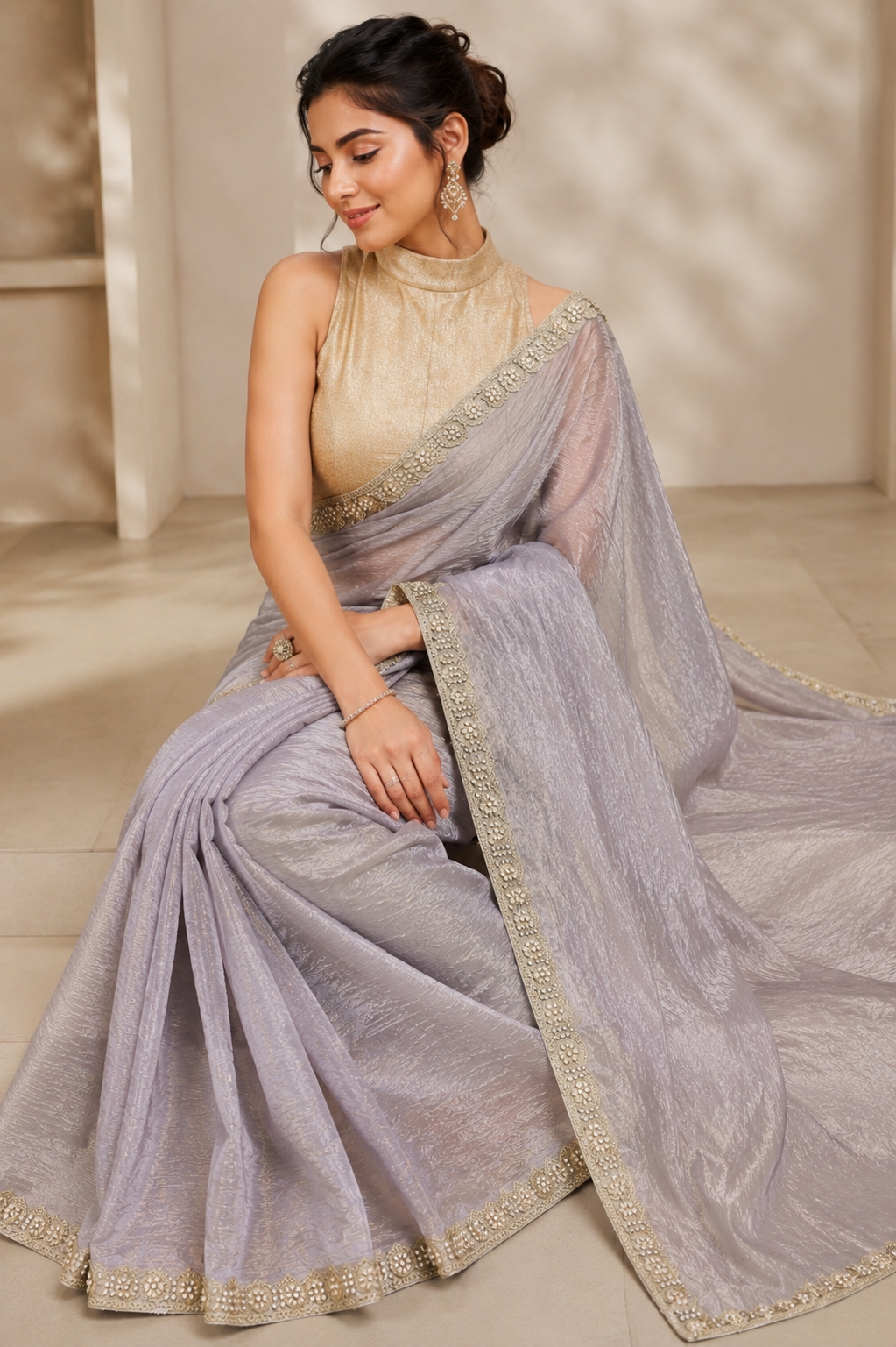 A woman seated indoors wearing a light lavender-gray saree with a beige sleeveless blouse and gold embroidered borders. The saree has a sheer dupatta draped over her shoulder and floor, with a soft neutral room background.