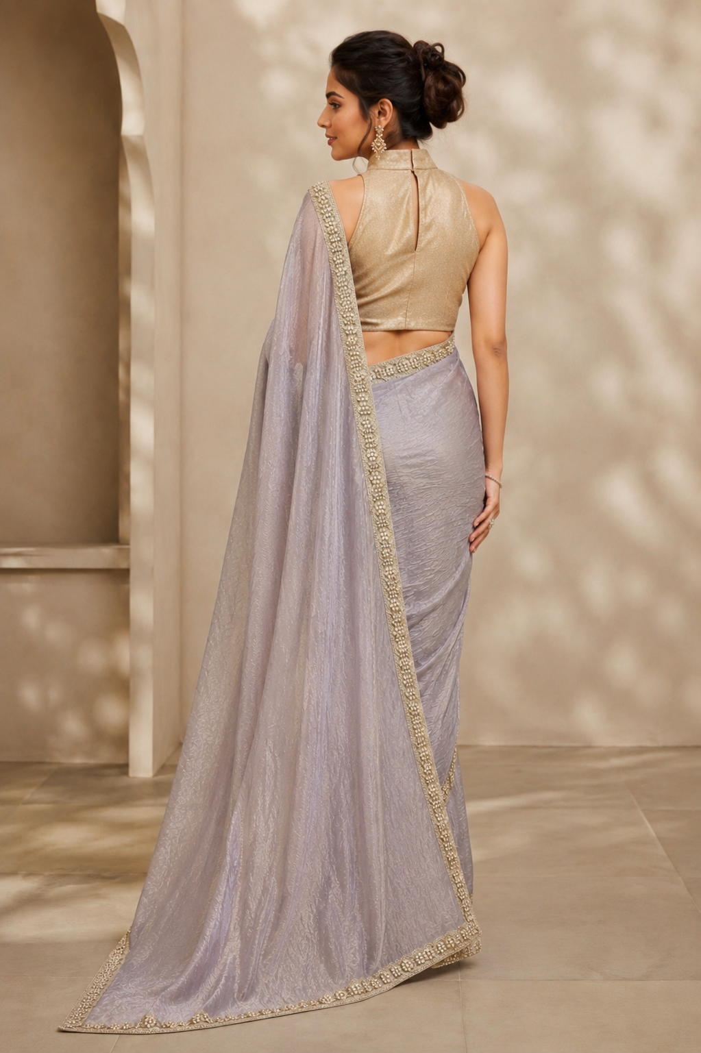 A woman shown from the back wearing a light lavender-gray saree with a beige embroidered blouse and a long matching dupatta, standing in a softly lit architectural setting.