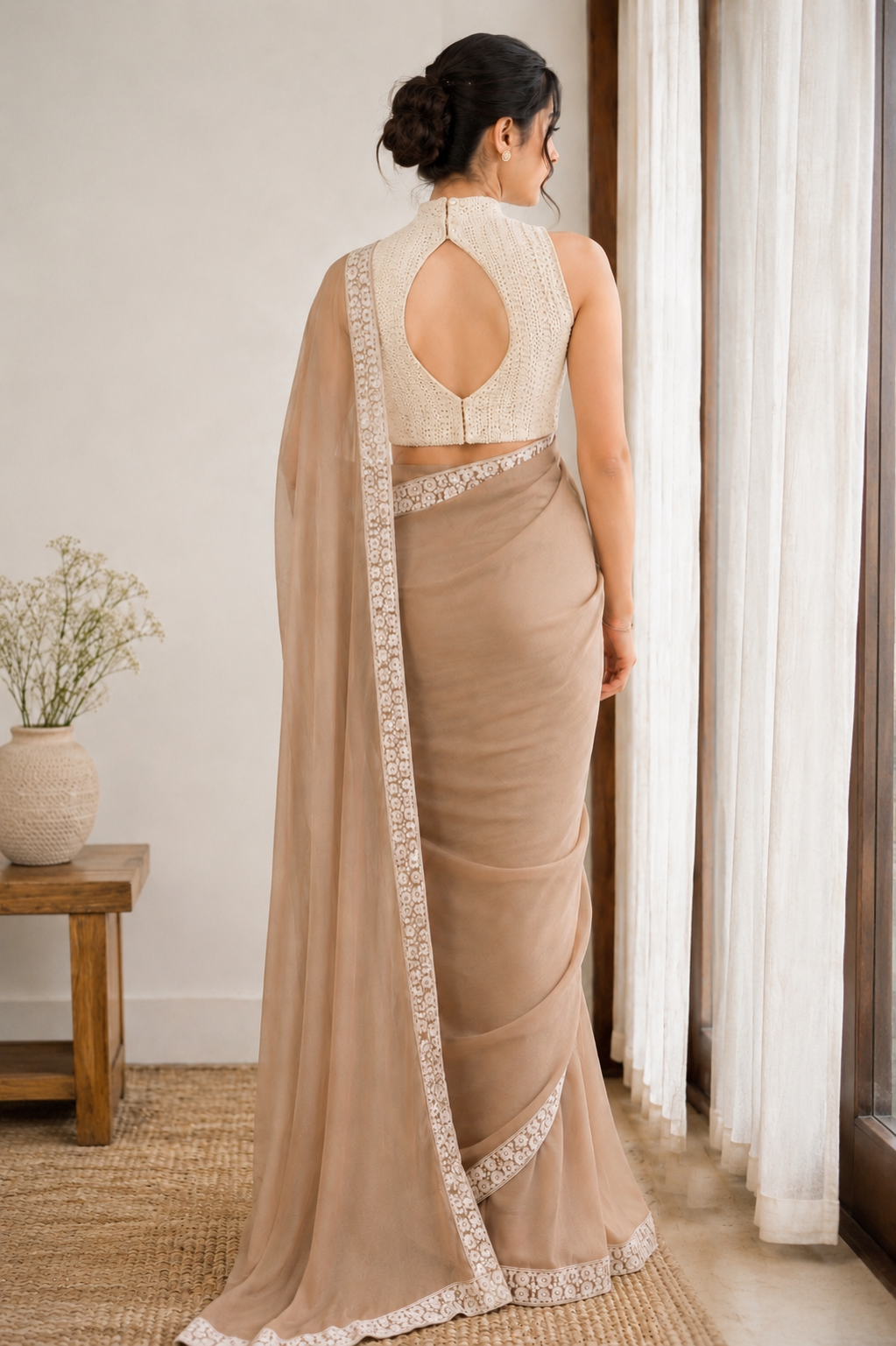 A woman stands with her back to the camera wearing a beige saree with a matching sleeveless blouse. The saree has white floral lace borders along the edge, and the blouse features a high neckline and an open back design.