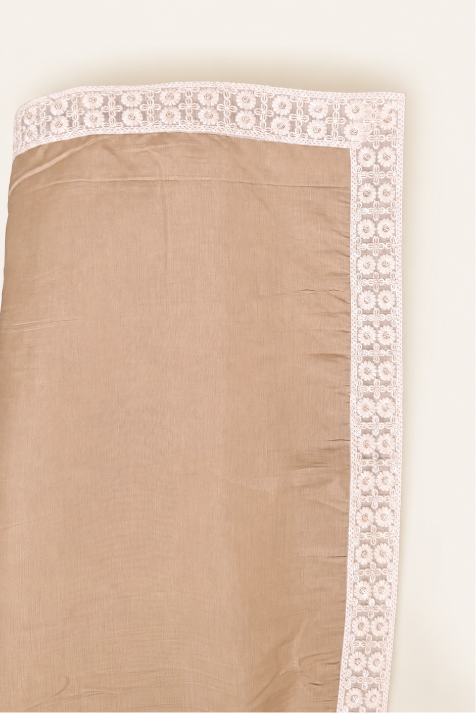 A close-up of a beige fabric item, likely a scarf or shawl, featuring a decorative white floral lace border along one edge and the bottom hem.