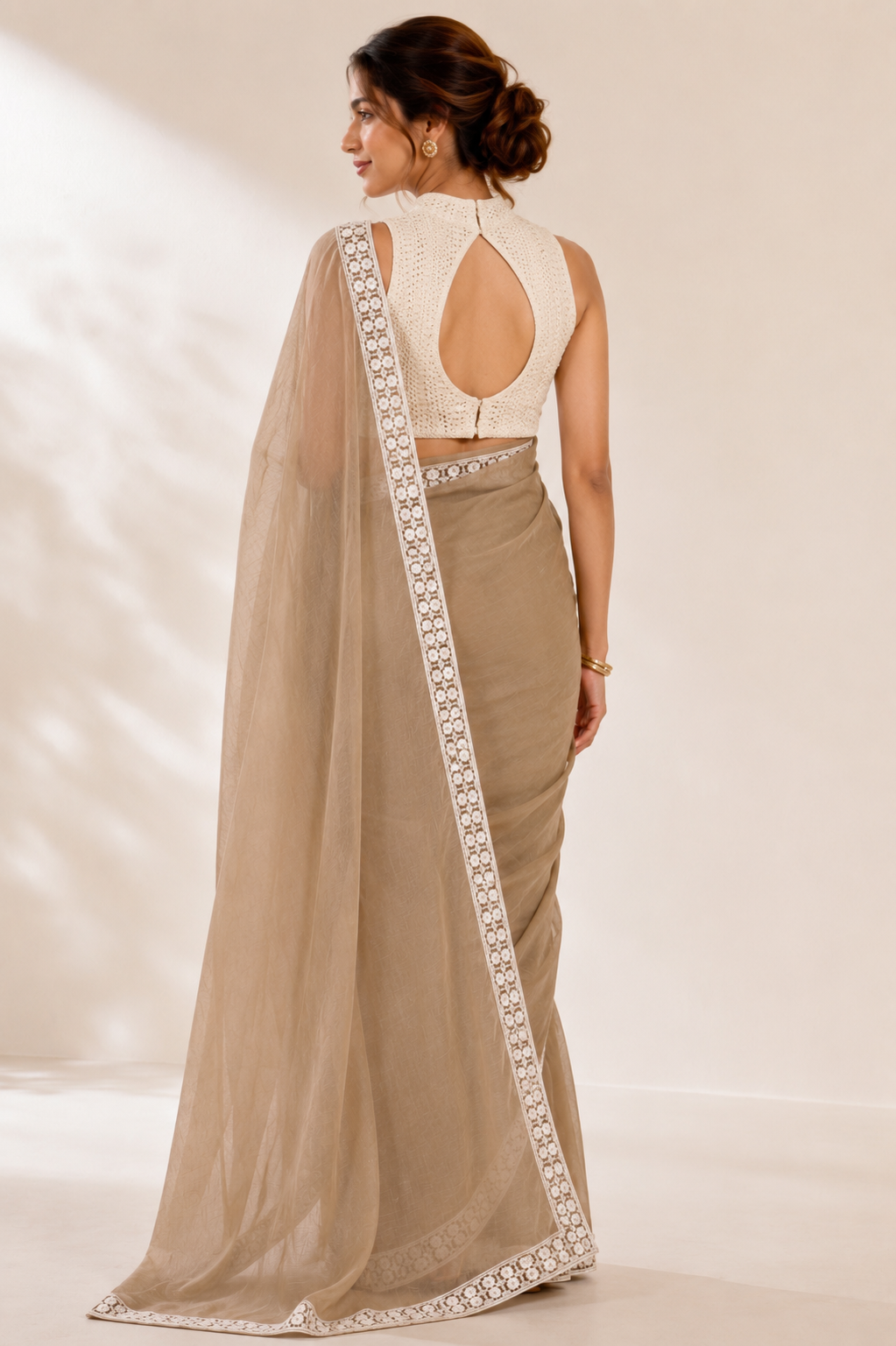 A woman shown from the back wearing a beige saree with a white embroidered border and a matching white lace open-back blouse, set against a warm beige studio background.