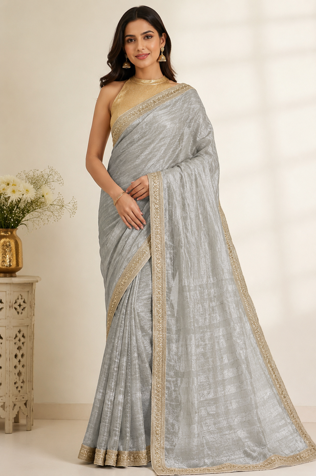 A woman modeling a light silver-gray saree with a gold border and matching gold blouse, standing in a softly lit indoor setting with a decorative side table and flowers at the side.