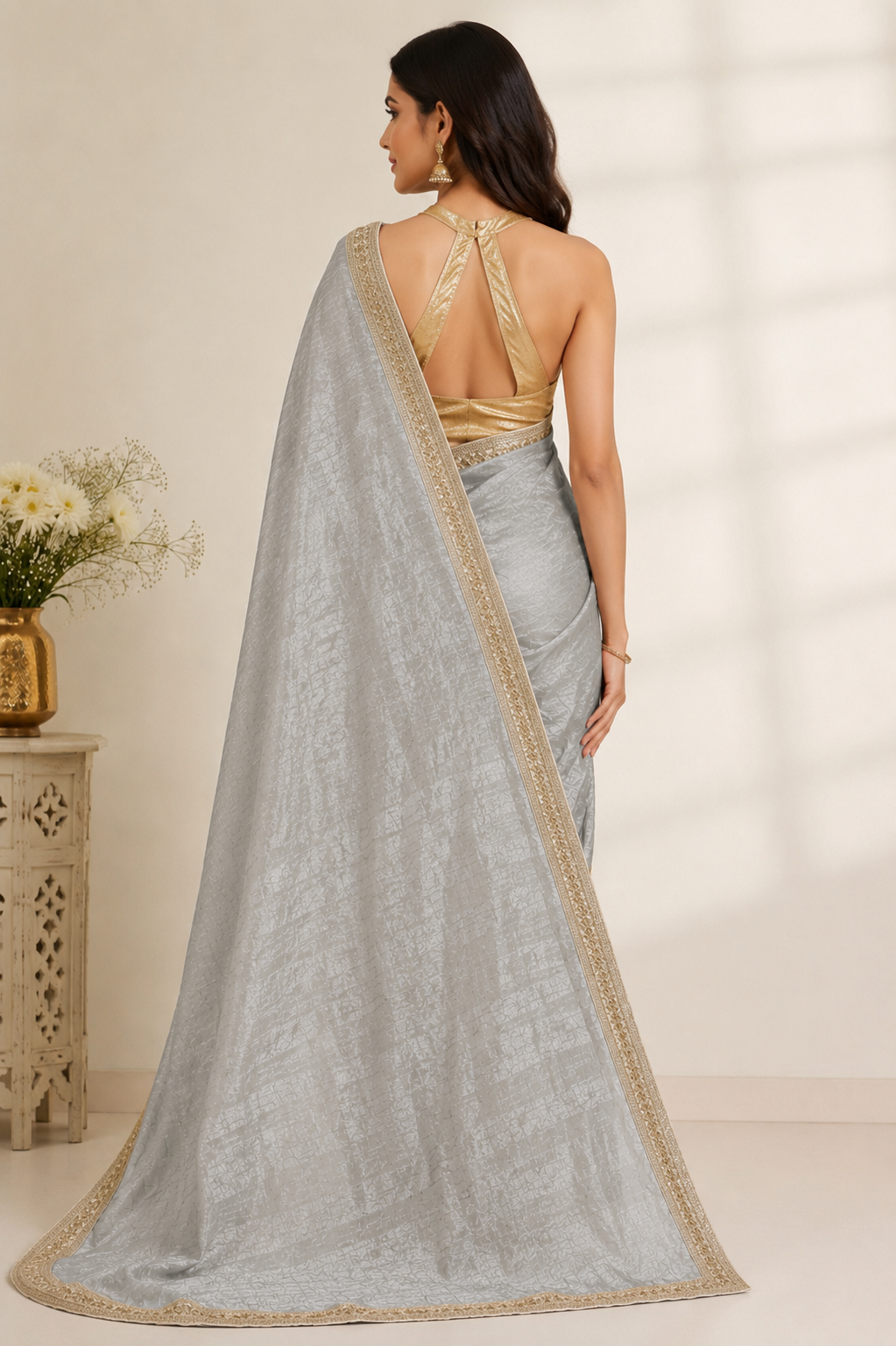 A woman shown from the back wearing a light silver-gray saree with a gold embroidered border and a matching gold embellished open-back blouse, standing in a softly lit indoor setting.