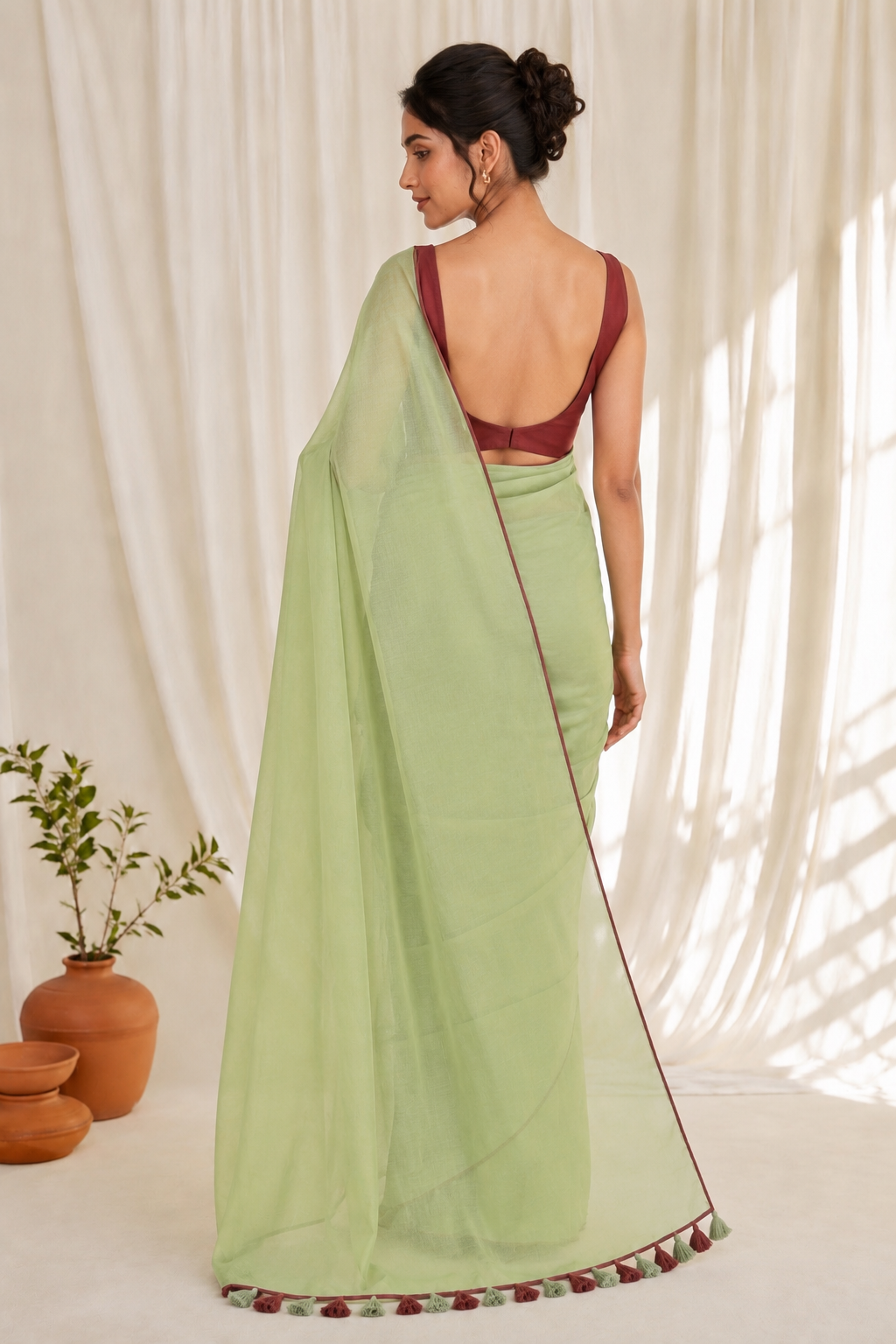 A woman wearing a light green saree with a deep maroon backless blouse, photographed from behind in a softly lit studio with cream drapes and terracotta pots.