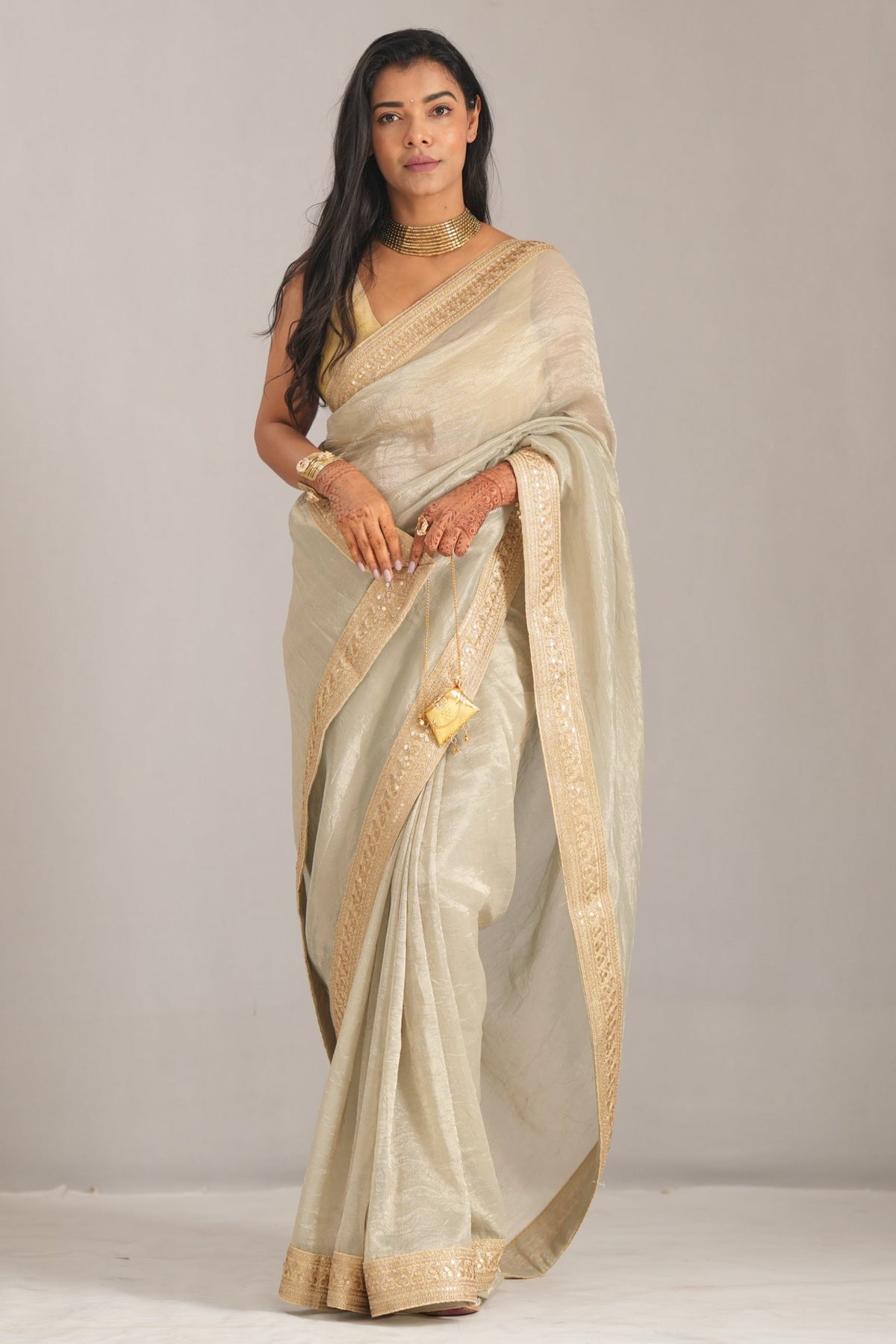 A woman modeling a light beige silk saree with a wide gold border, standing against a neutral light gray studio backdrop.