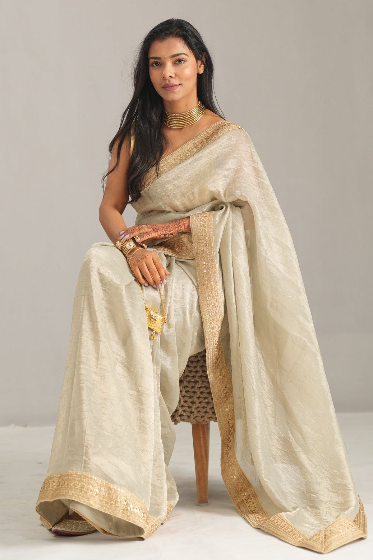A woman seated on a small stool wearing a draped beige saree with a gold border, photographed against a plain light gray studio background.