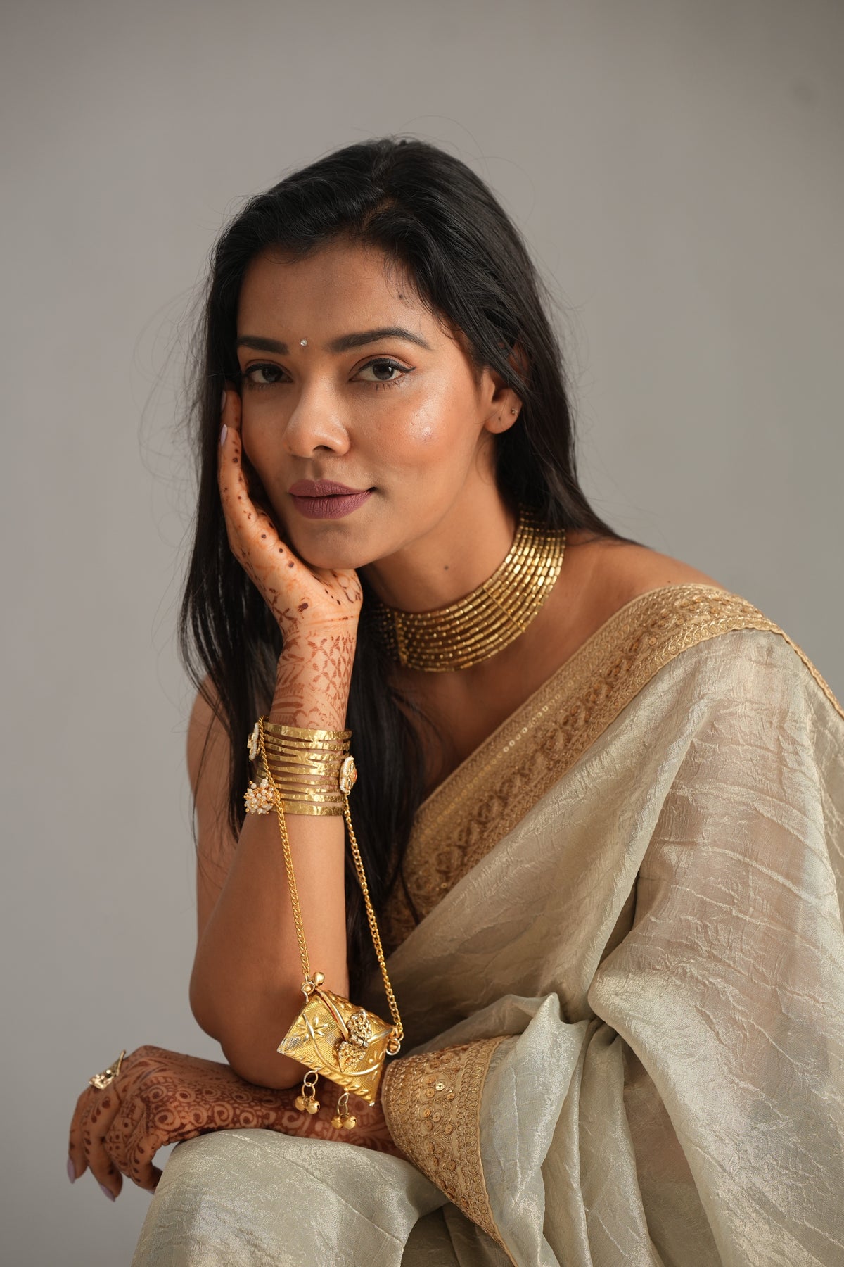 A woman wearing a beige and gold saree with traditional Indian jewelry, including a gold necklace, bangles, and a pendant, posing against a neutral gray background.