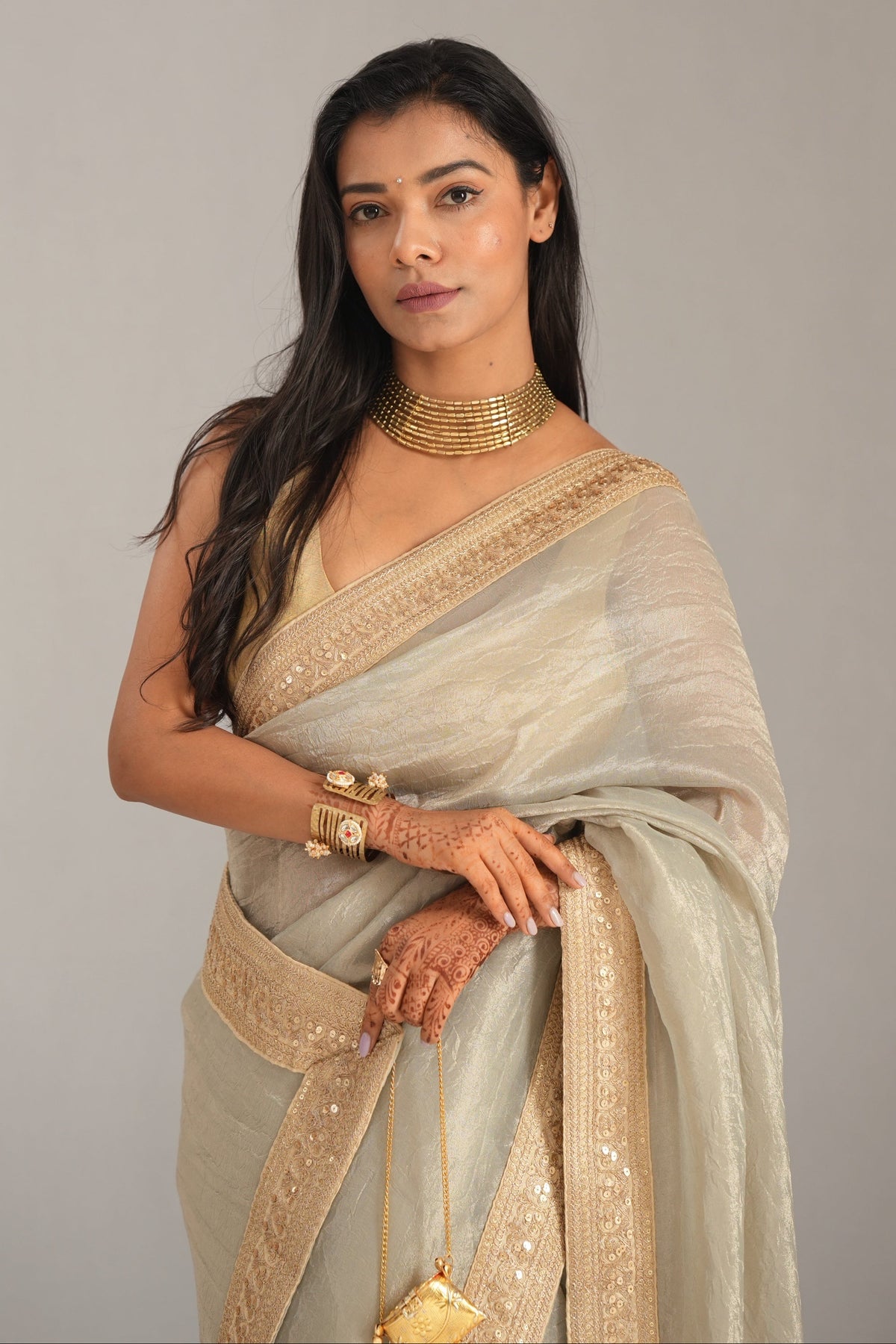 A woman modeling a light beige and gold saree with ornate gold jewelry against a plain gray studio background.