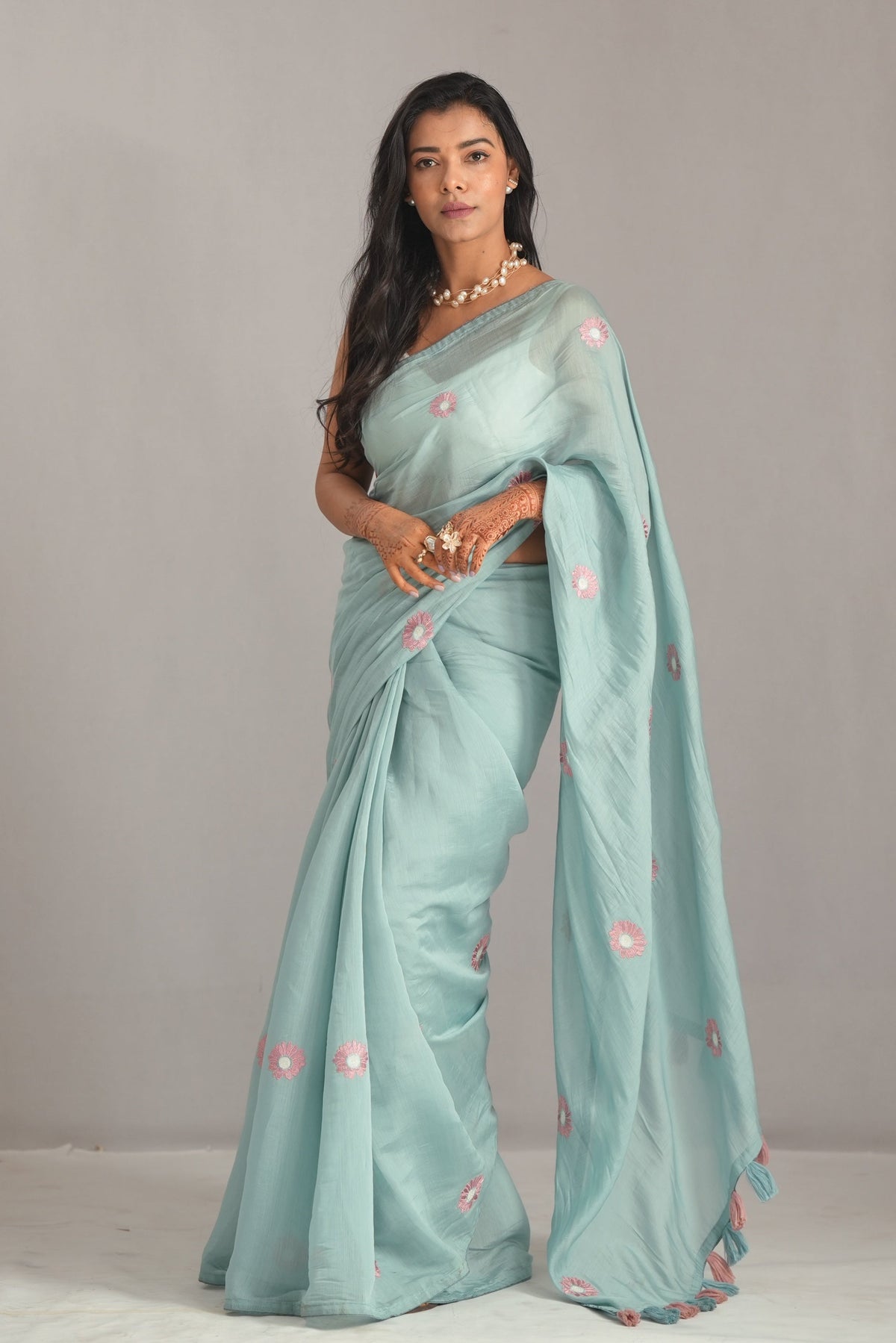 A woman modeling a light blue-green saree with pink floral embroidery, standing against a neutral gray studio backdrop.
