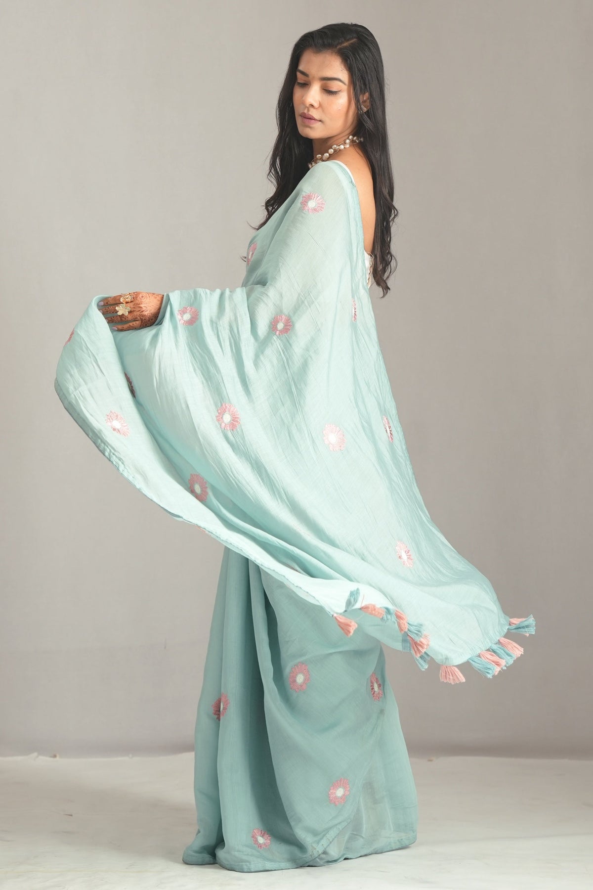 A woman modeling a flowing mint green saree with pink floral embroidery, posed against a neutral gray studio backdrop.