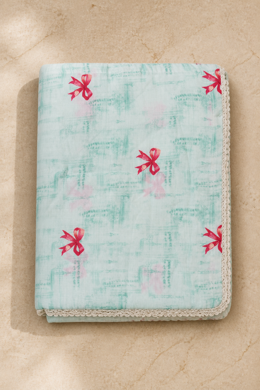 A folded fabric item, likely a blanket or throw, featuring a light aqua and pale pink abstract pattern with small red bow motifs and a cream lace trim, displayed on a beige stone surface.