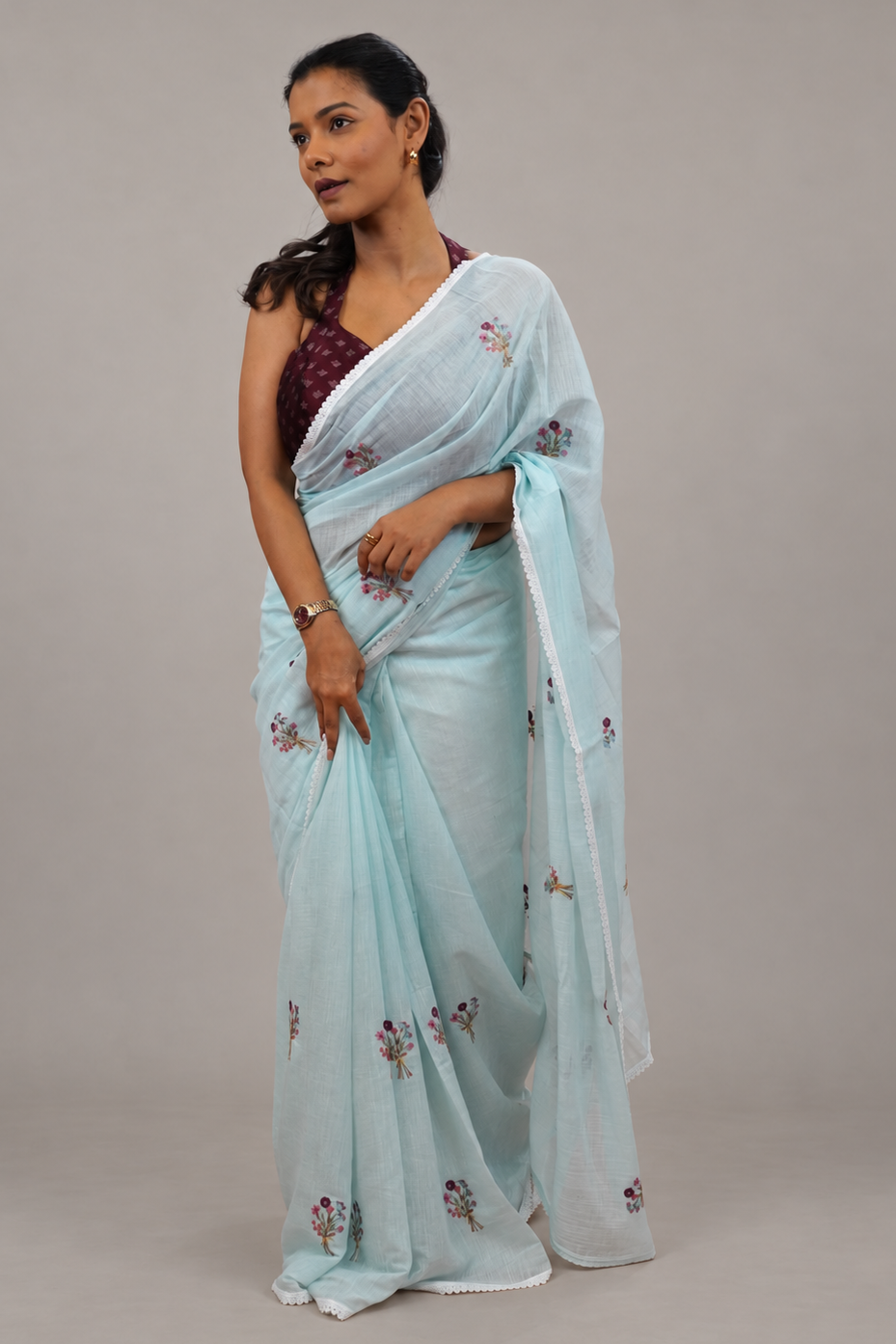 A woman modeling a light blue semi-sheer saree with small floral embroidery, paired with a dark purple patterned blouse, against a solid gray studio background.