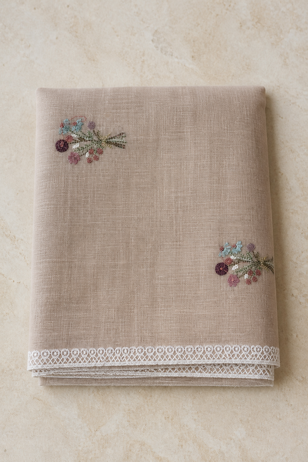 A folded beige linen tablecloth with delicate floral embroidery and a white lace border, displayed on a light beige stone surface.