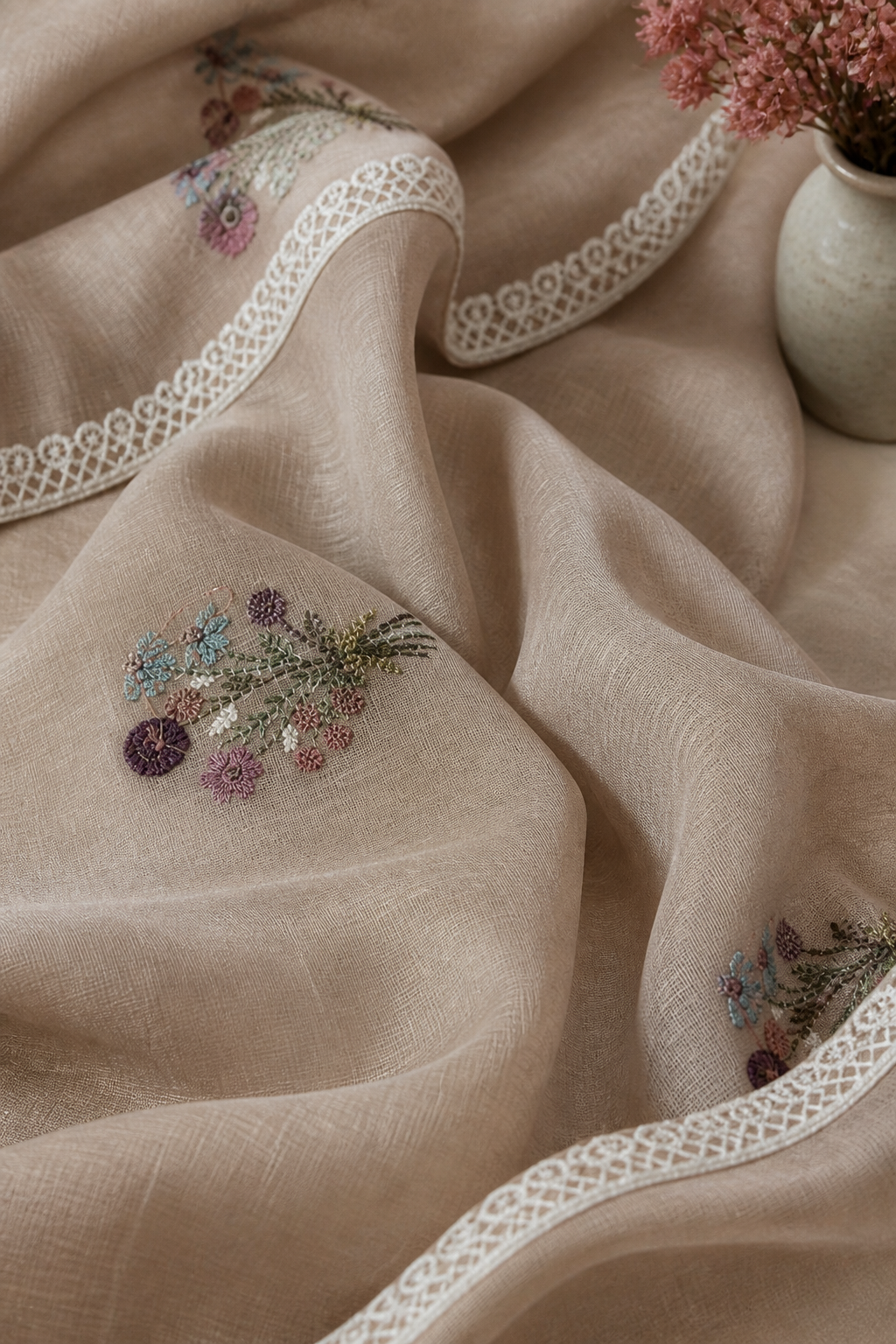 A draped beige linen fabric with delicate white lace trim and embroidered floral motifs in purple, blue, and green, styled with a small vase of dried pink flowers.