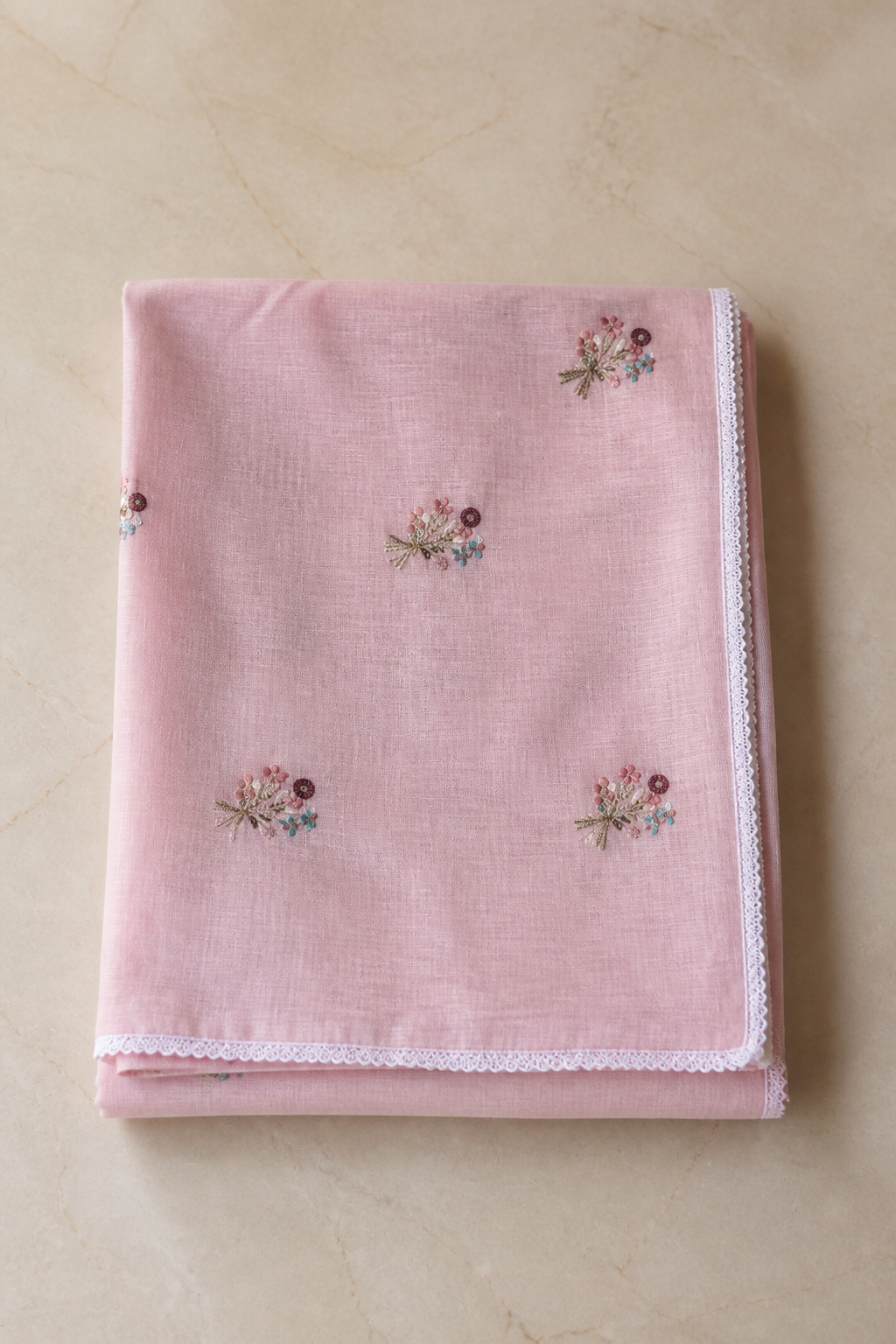 A folded light pink fabric item, likely a baby blanket or cloth napkin, featuring small embroidered floral motifs and a white lace trim along the edge, displayed on a beige marble surface.