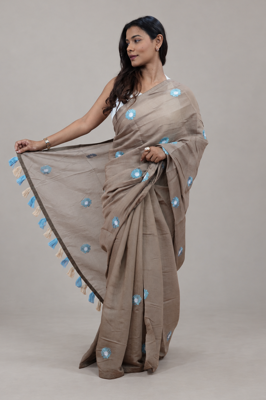 A woman modeling a taupe-beige cotton saree with light blue floral motifs and a matching border, posed against a neutral gray studio background.