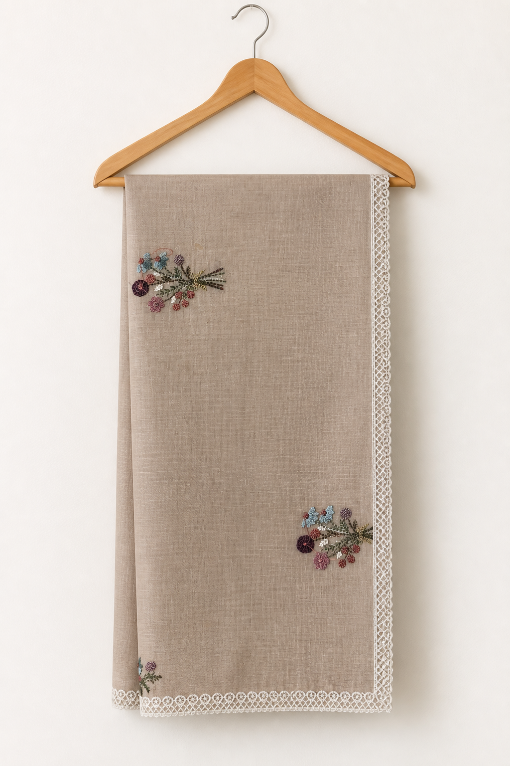 A beige linen tablecloth with white lace trim and embroidered floral details, hanging on a wooden hanger against a plain off-white wall.