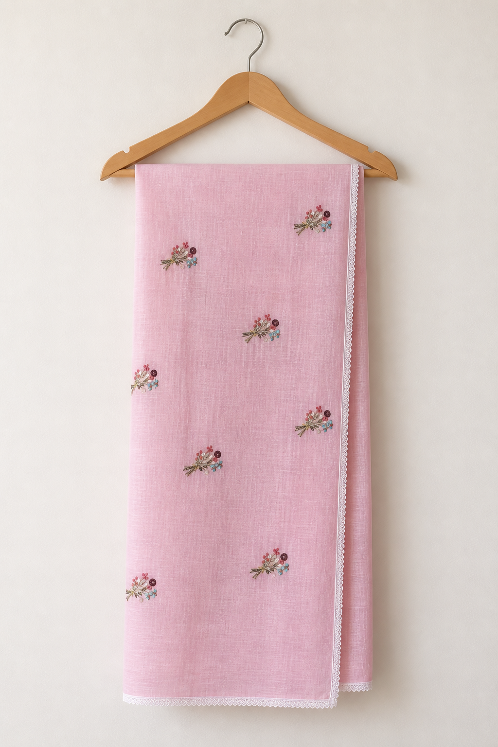 A light pink cotton muslin scarf with small embroidered floral motifs, hanging on a wooden hanger against a plain off-white wall.