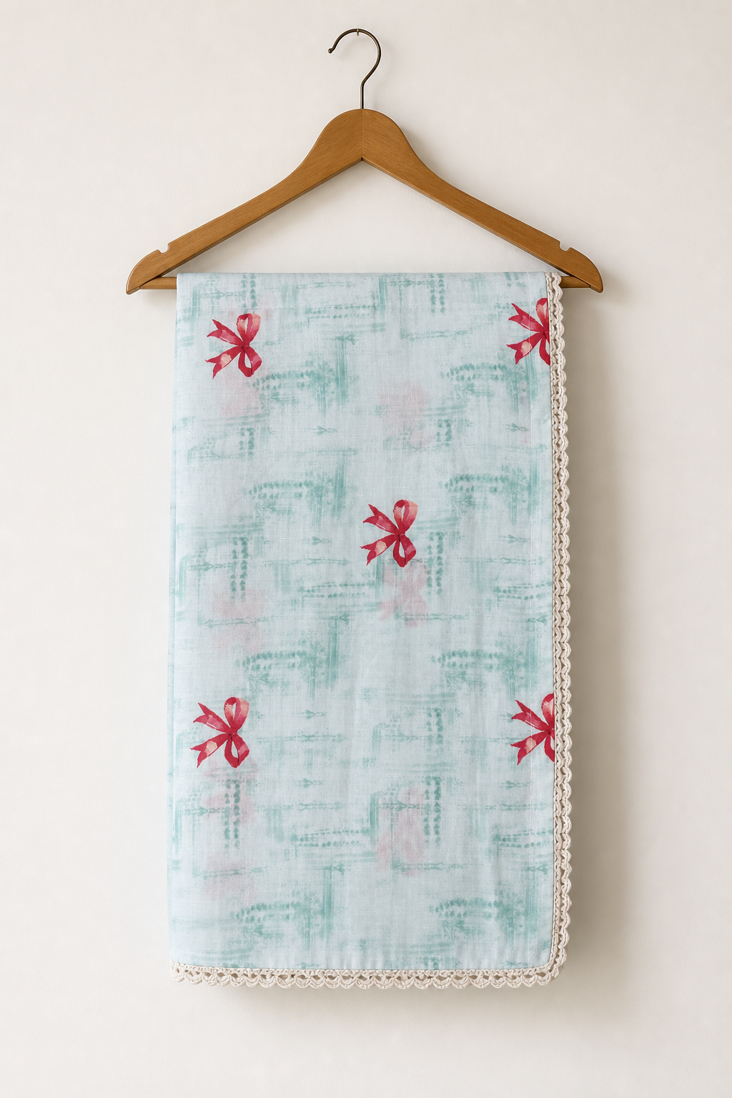 A light blue and white patterned cotton blanket with red bow motifs and a cream lace trim, hanging from a wooden hanger against a plain off-white wall.