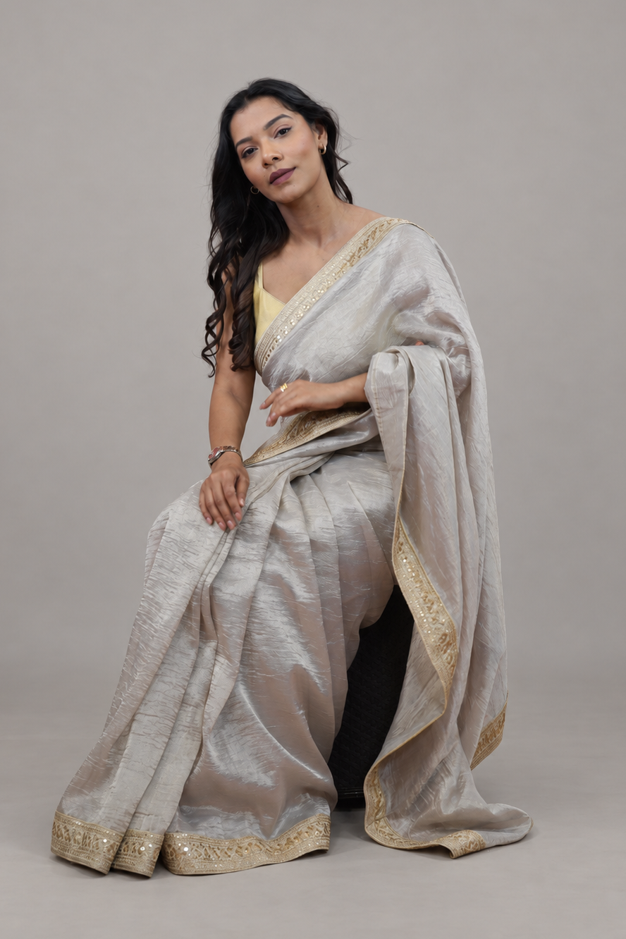 A woman models a light silver-gray silk saree with gold zari borders, paired with a yellow blouse, seated against a solid gray studio background.