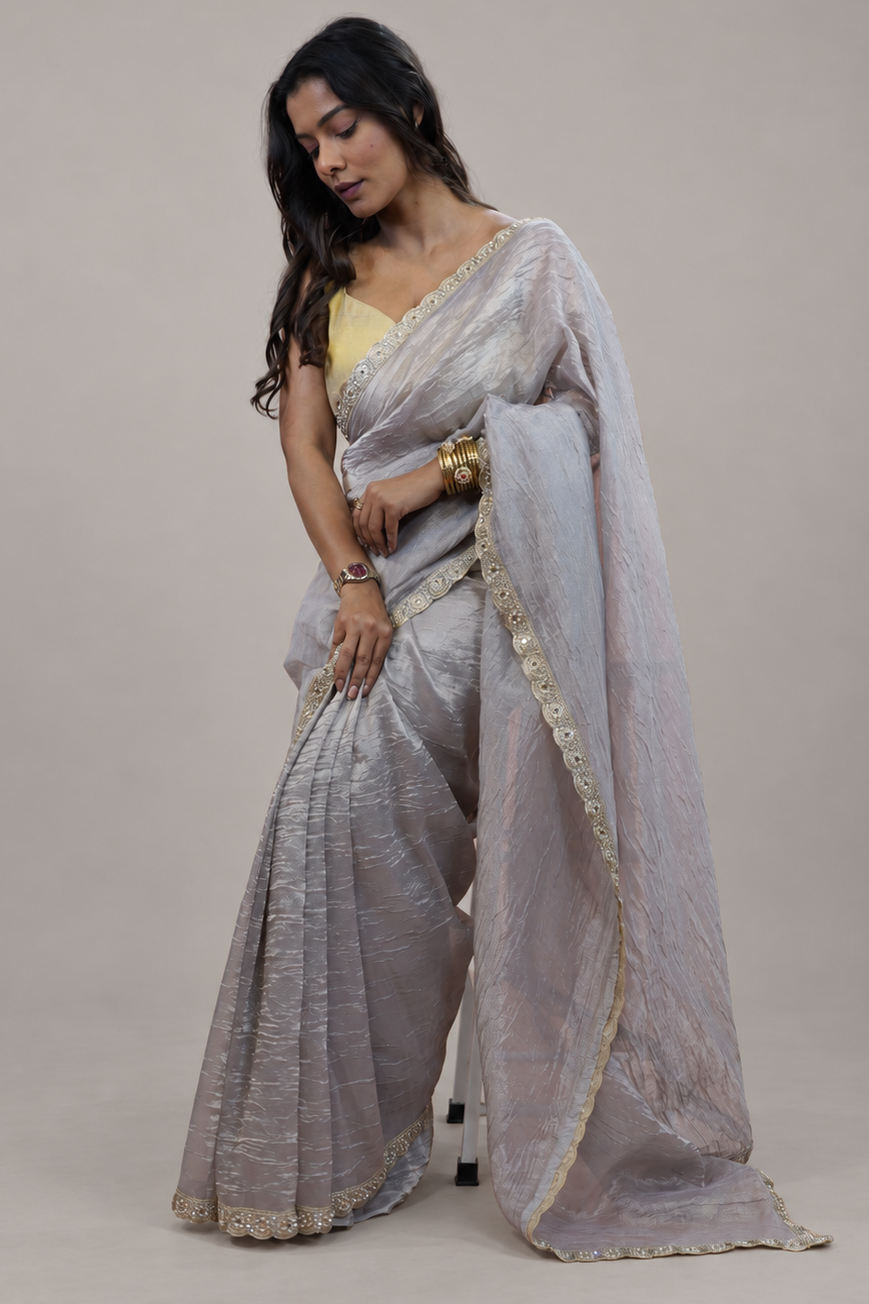 A woman modeling a light silver-gray saree with gold embroidered borders and a yellow blouse, standing against a plain beige studio background.